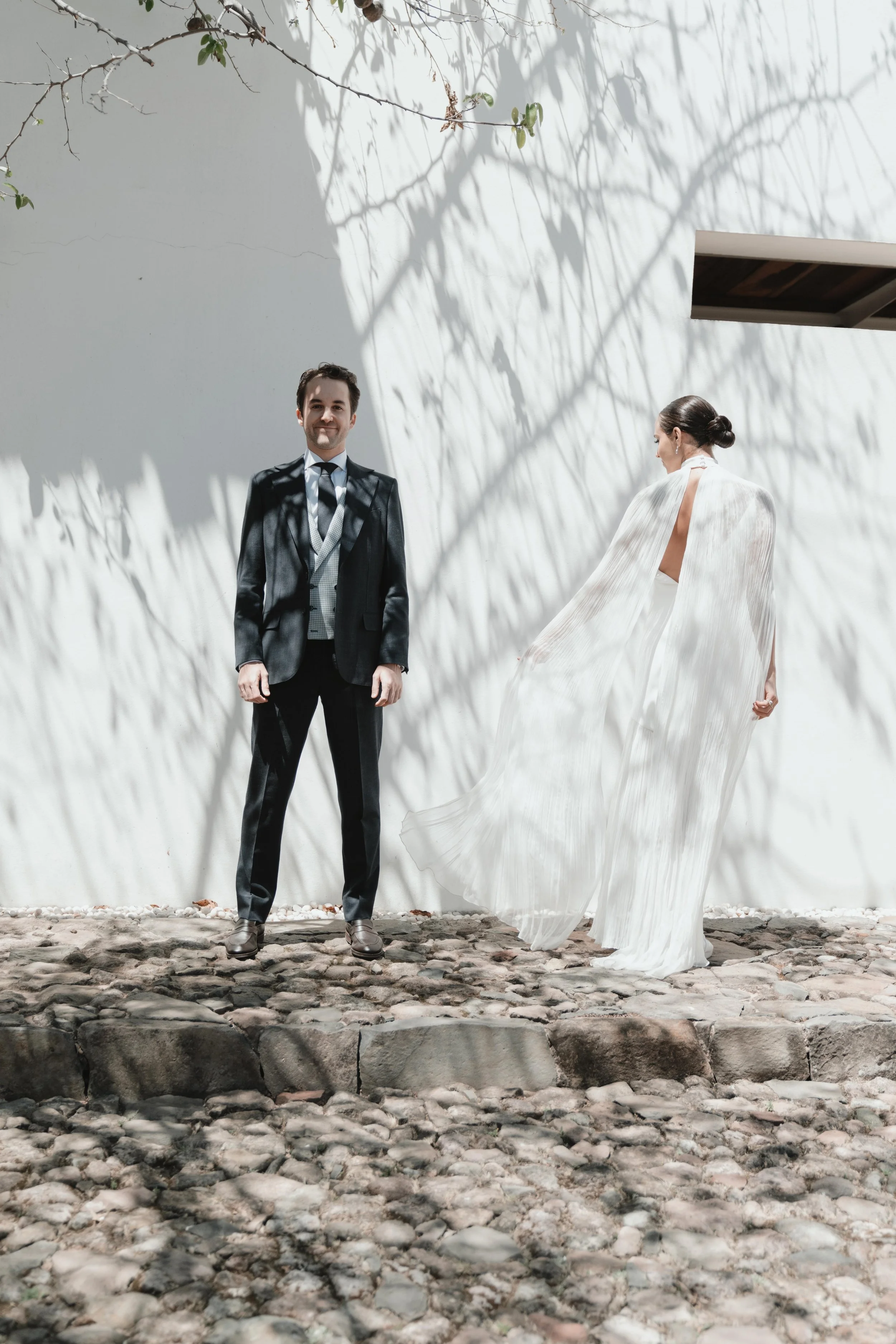 Pareja en ropa formal de boda, el hombre con traje oscuro y la mujer con vestido largo y fluido blanco en un escenario exterior con sombra de ramas en la pared.