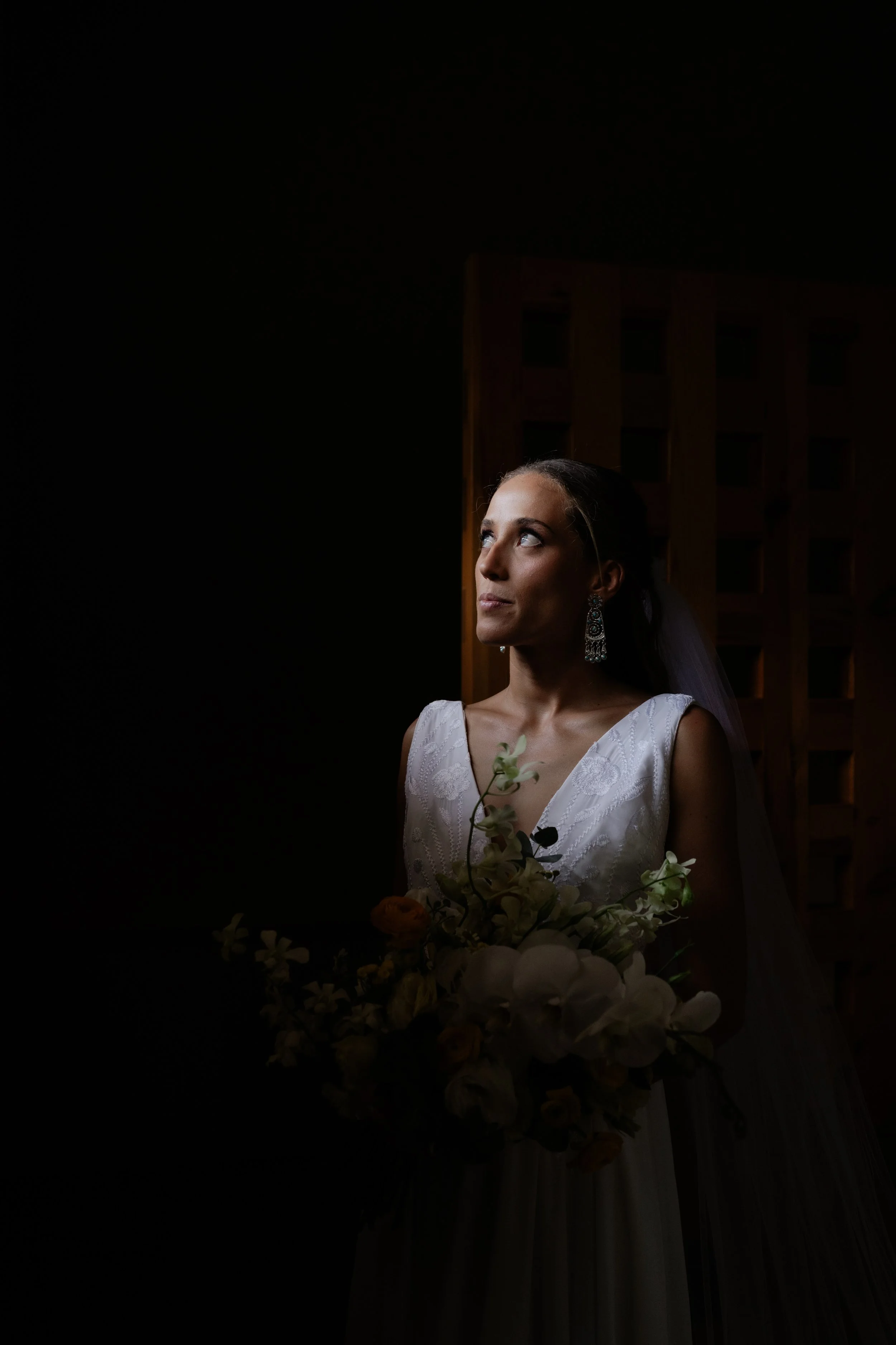 Mujer vestida de novia con pendientes elegantes, sosteniendo un ramo de flores, en un ambiente oscuro con luz suave en su rostro.