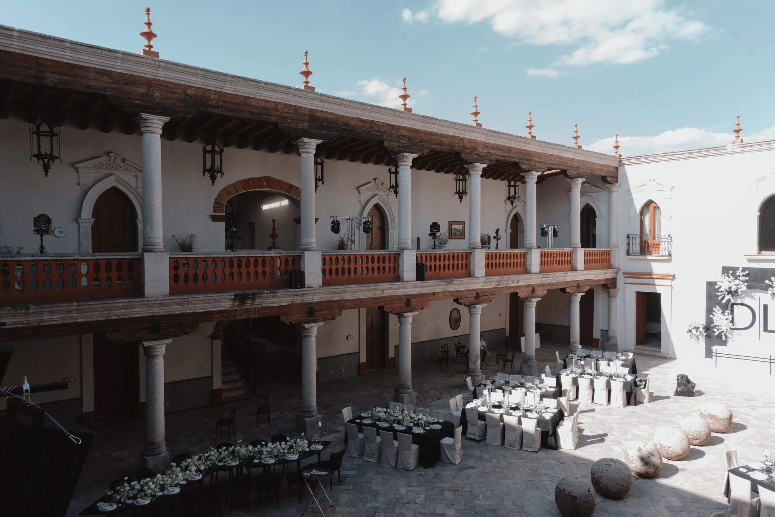 Un patio interior de estilo colonial con columnas de piedra y un balcón de madera, decorado para una celebración con mesas arregladas con manteles blancos y centros de flores, y sillas con fundas blancas, en un día soleado con el cielo despejado.