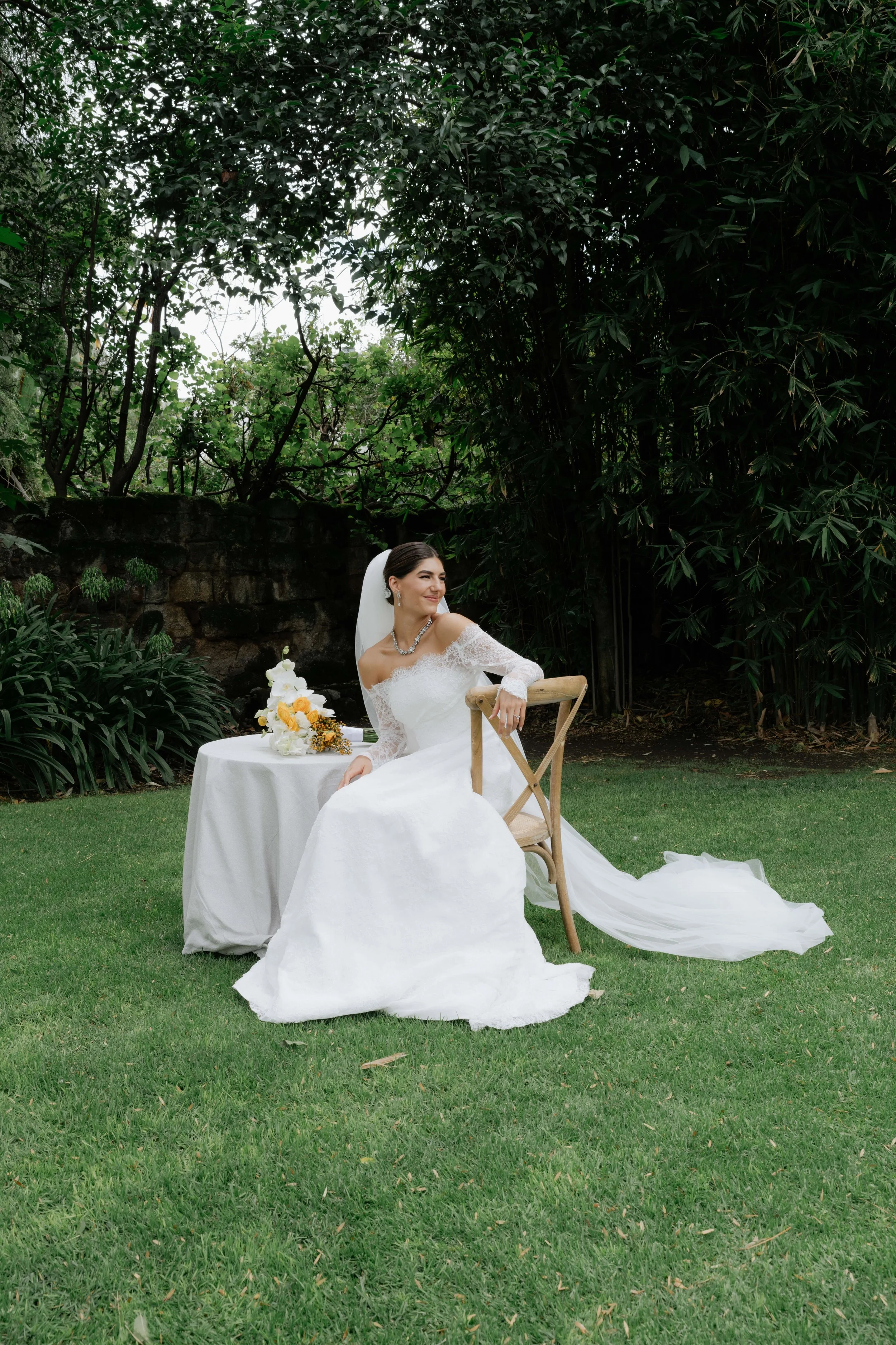 Una novia vestida de boda blanca con vestido de encaje y velo, sentada en una silla en un jardín con césped verde y plantas frondosas, junto a una mesa con flores.