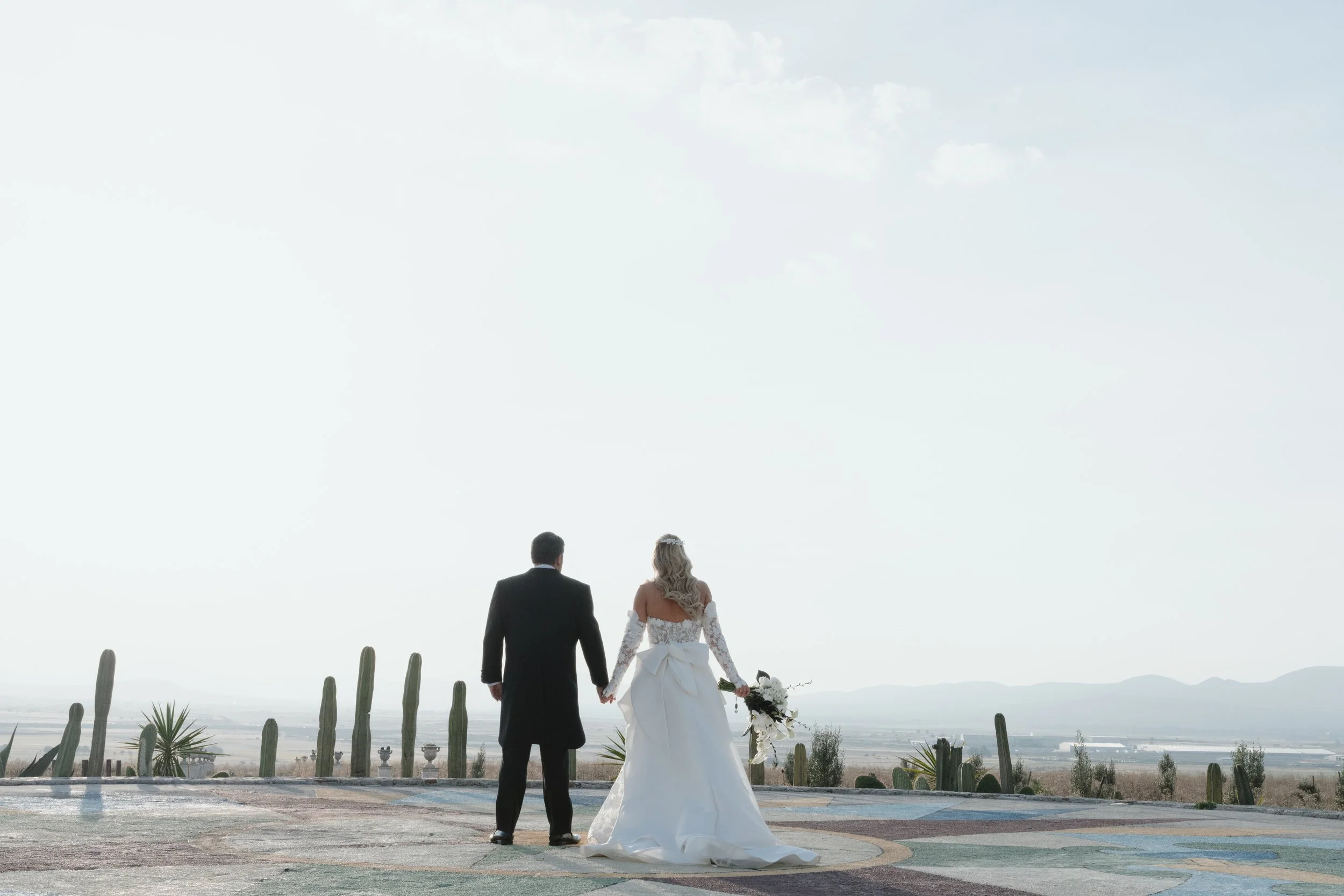 Pareja de novios caminando juntos en un paisaje abierto con cactus y montañas en el fondo, en un día soleado.