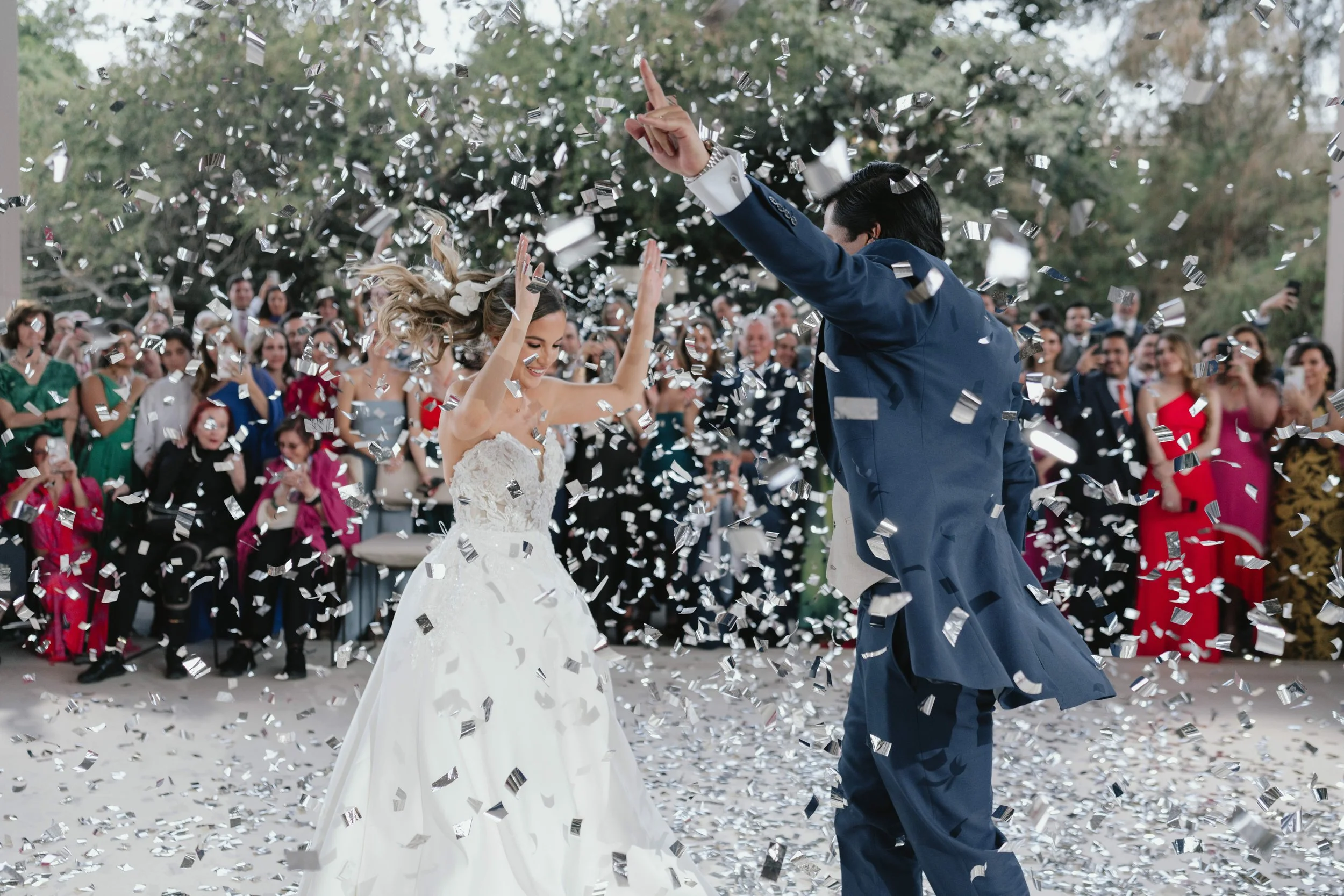 Una pareja de novios bailando entre confeti en una boda, con invitados en el fondo