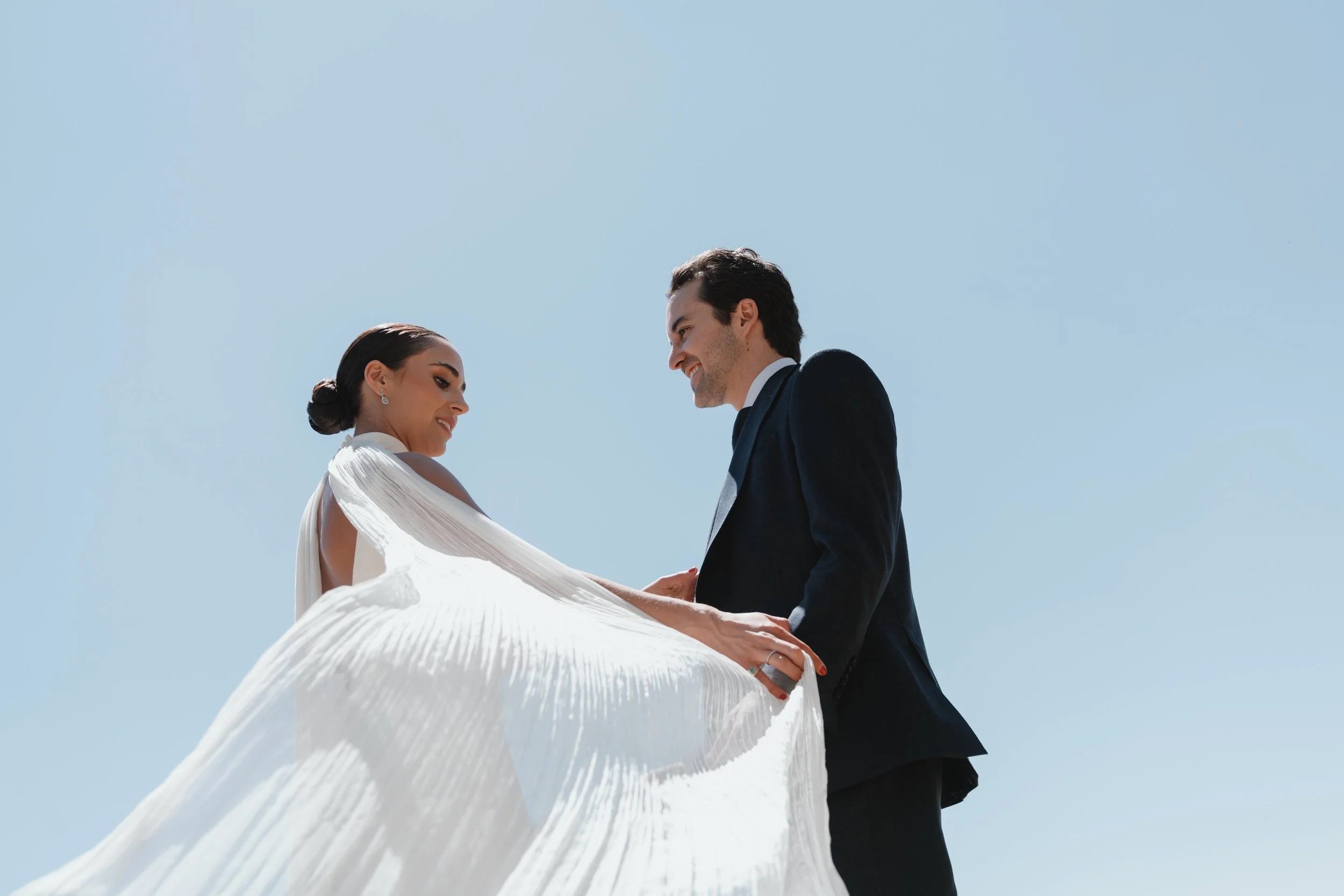 Pareja de recién casados mirándose con cielo azul de fondo, ella en vestido de novia y él en traje formal.