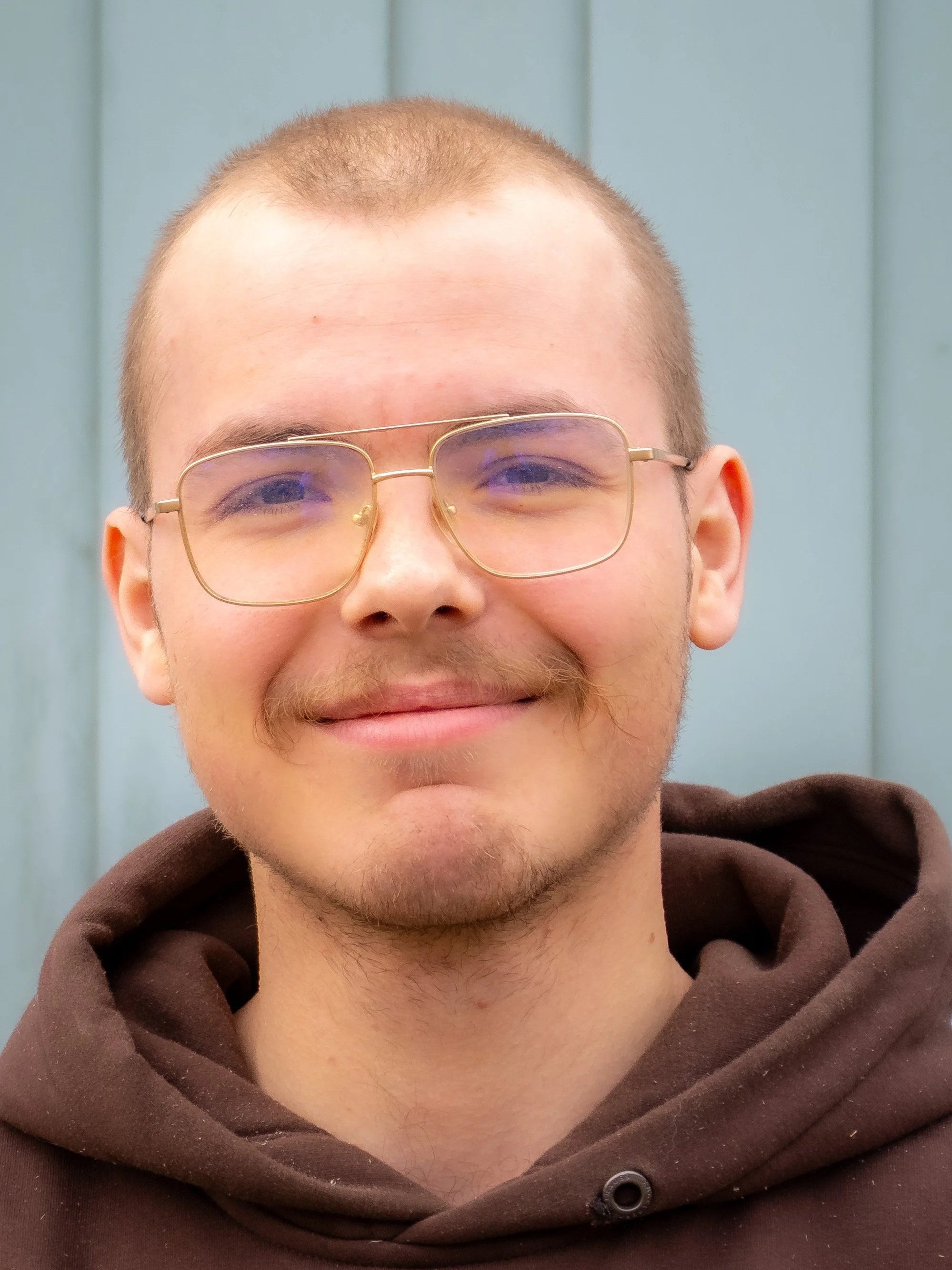 A young man with glasses, a slight smile, and a beard, wearing a brown hoodie, standing in front of a pale blue wooden wall.