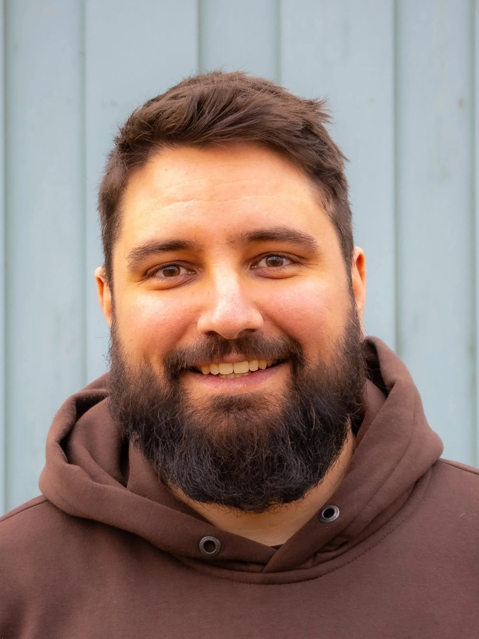 Close-up of a smiling man with a beard and short brown hair, wearing a brown hoodie, standing in front of a light blue wooden wall.