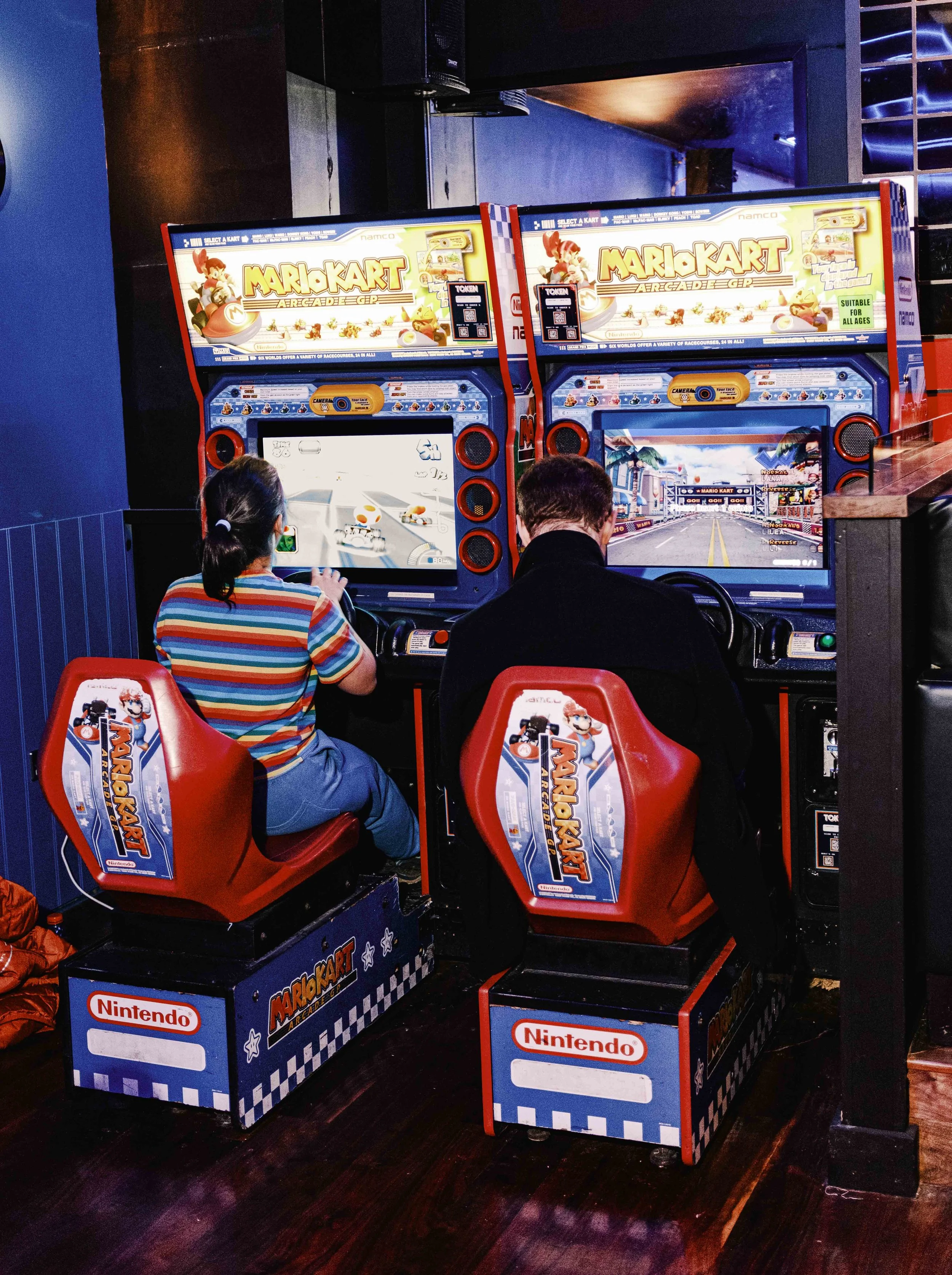 Two people playing Mario Kart arcade game in an arcade, sitting in red racing seats with Nintendo branding, with game screens displaying racing tracks.