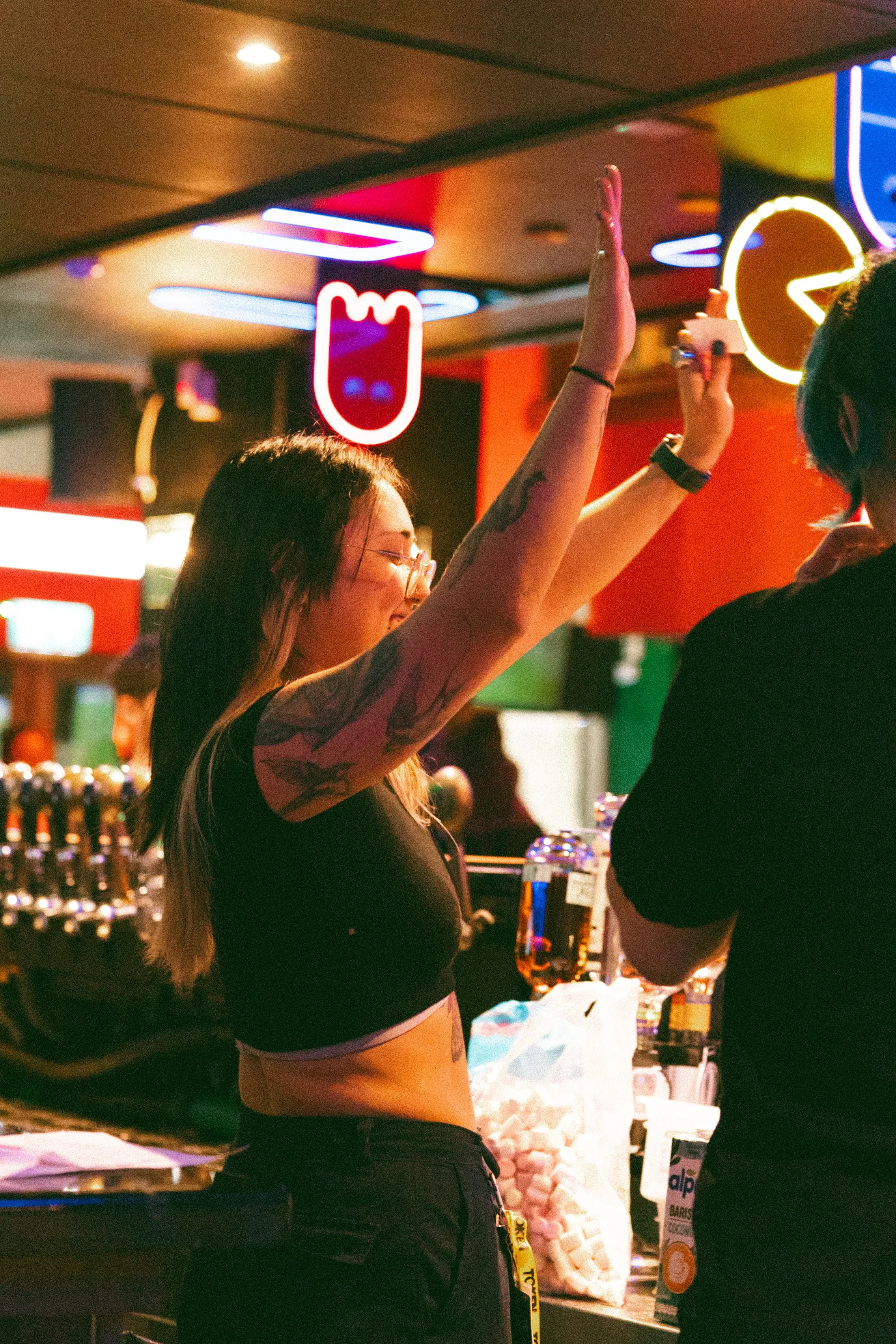 A woman with tattoos on her arms and glasses is raising her arm in a lively bar or nightclub, surrounded by colorful neon lights and other patrons.