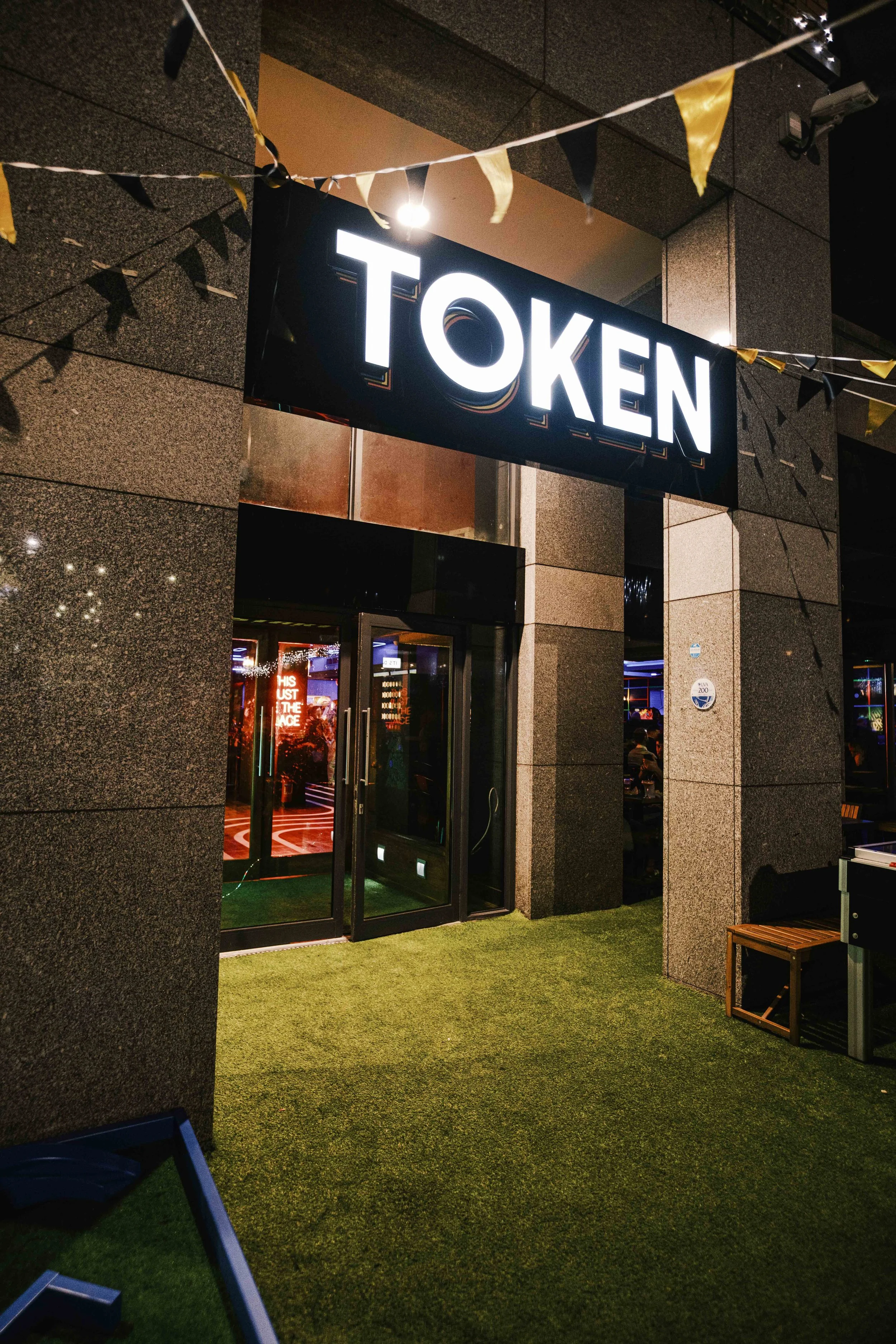 Night view of the entrance to a venue or club with a large illuminated sign that says 'TOKEN'. There are decorative yellow and black flags hanging above the entrance, and the doorway is open, revealing a glimpse of the interior with colorful lights.