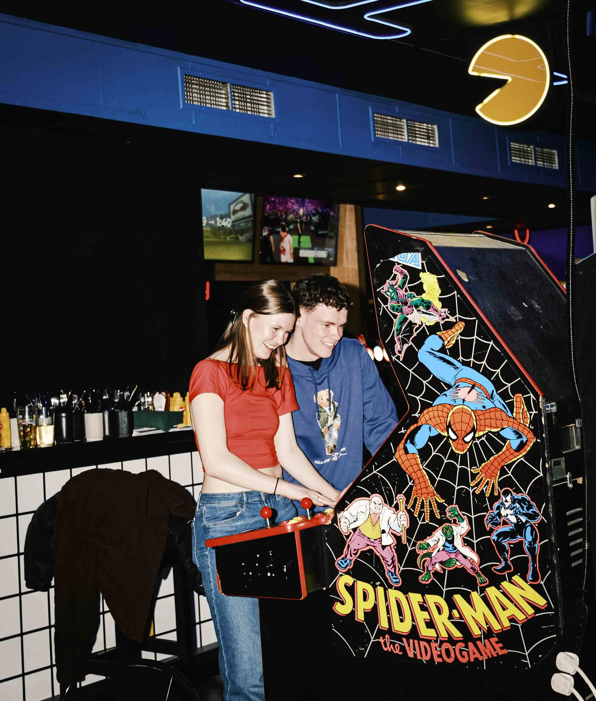 Two young people, a woman and a man, are playing a Spider-Man themed arcade game in a dimly lit arcade or gaming venue, both smiling and enjoying the game.