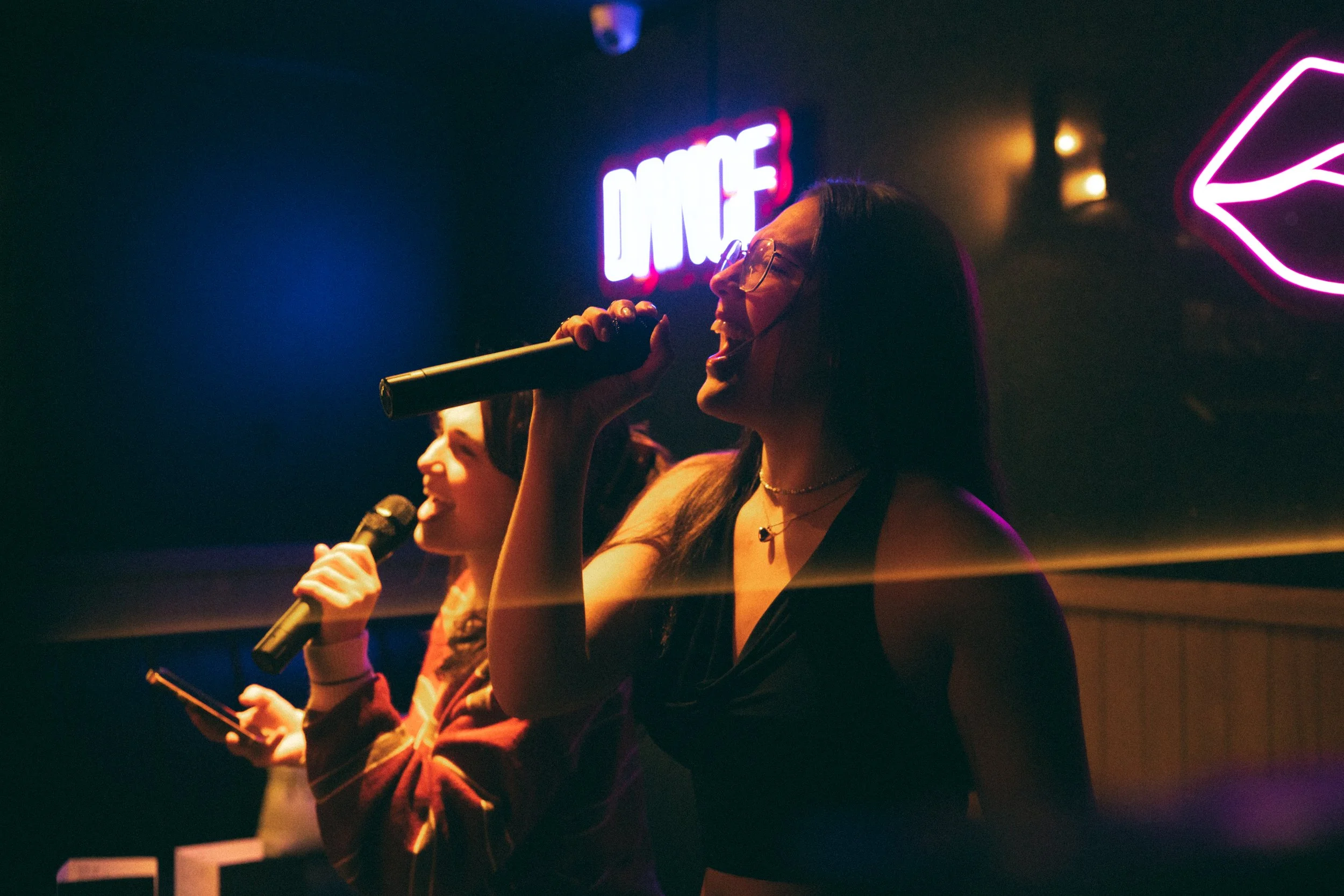 Two women singing into microphones in a dimly lit karaoke bar with neon signs, one of which says "DANCE" and the other is lips.