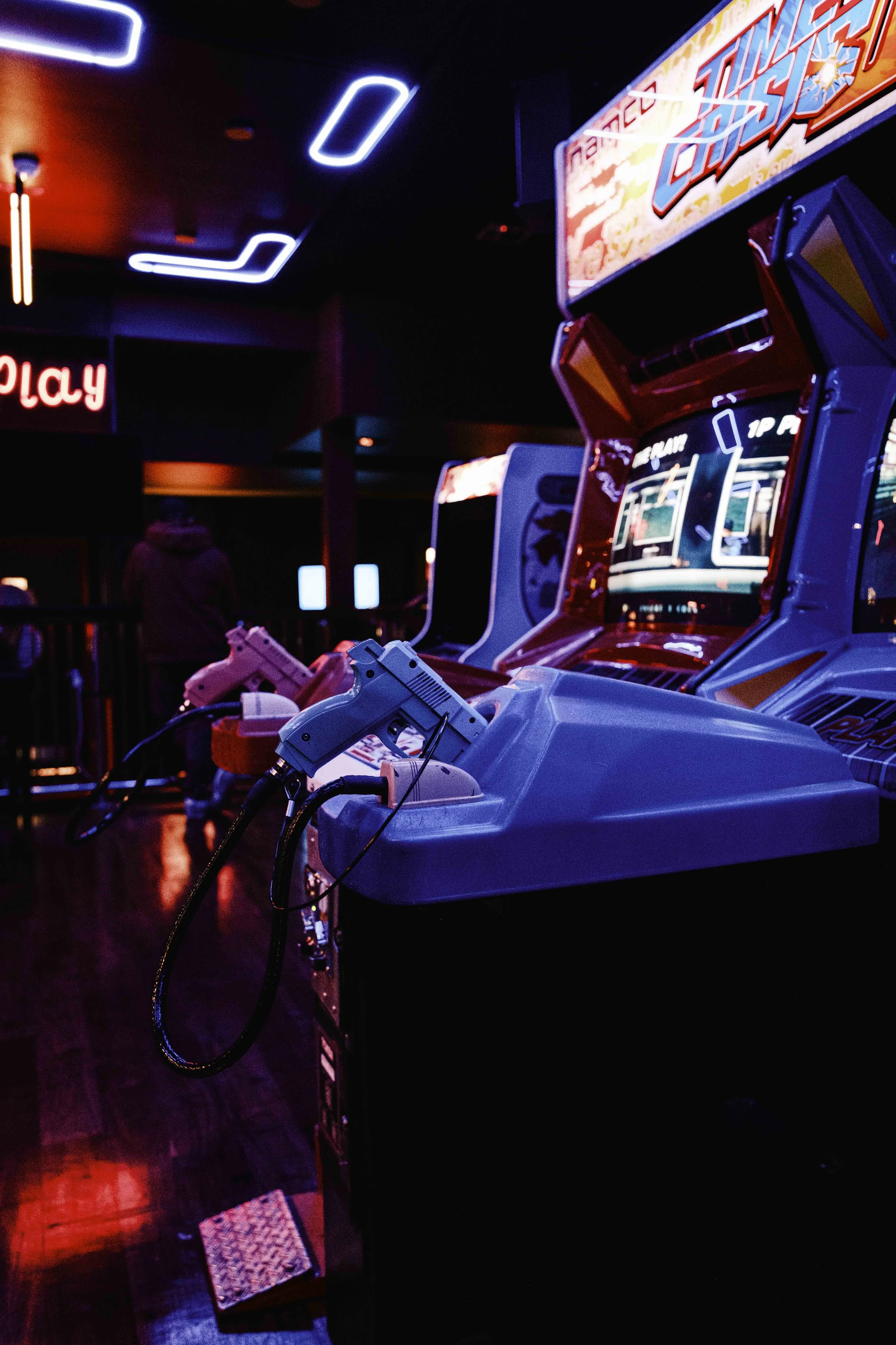 Arcade game machines withguns at a gaming arcade, illuminated by neon signs, in a dark room with some people in the background.