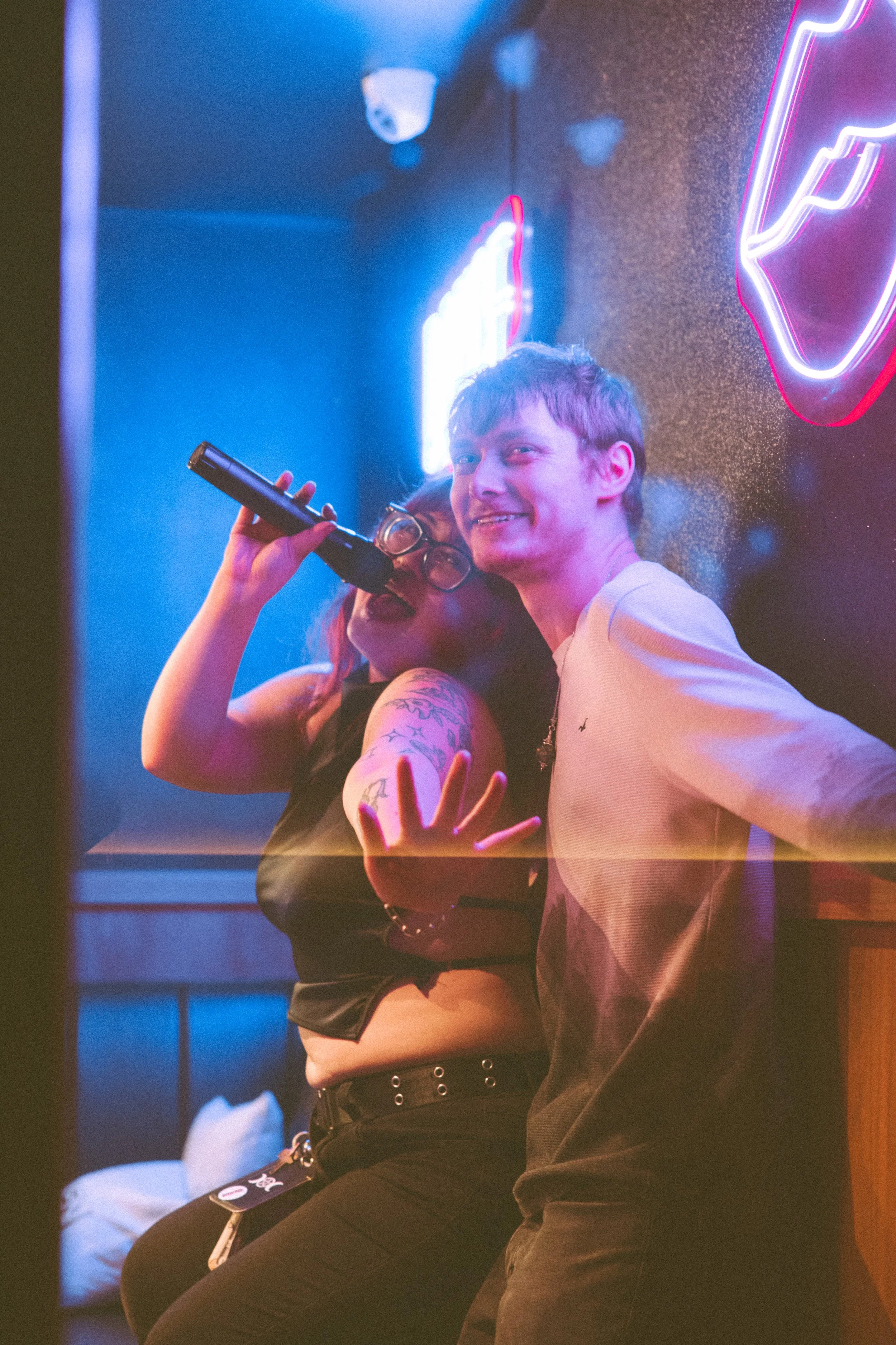 Two people singing karaoke in a dimly lit room with blue and pink neon lights, one holding a microphone, the other smiling.