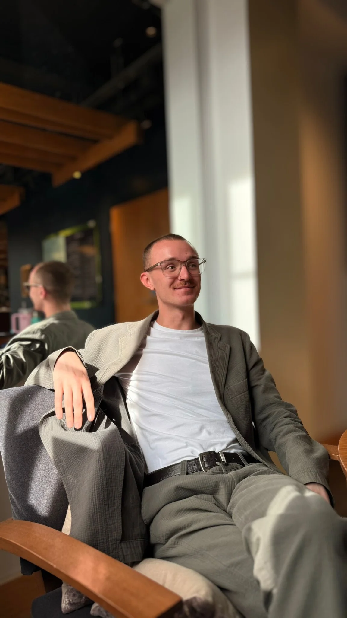 A man with glasses, a short fringe, and a mustache is sitting and leaning back comfortably in a chair, smiling slightly. He is wearing a light-colored suit jacket, a plain white T-shirt, and gray pants, with a black belt. Behind him, there is a mirror reflecting his side profile and the ambient interior of a room with warm lighting, wooden accents, and a hanging ceiling structure.