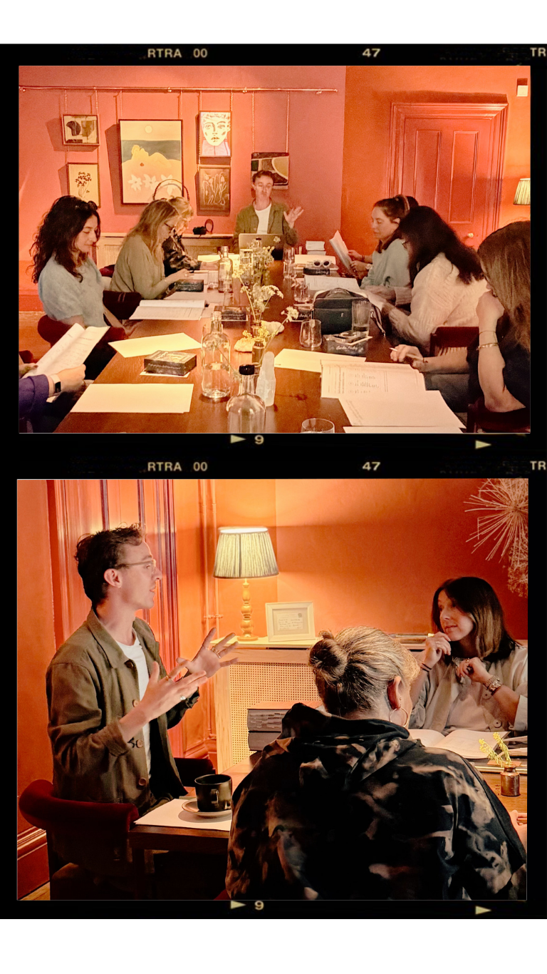 A group of people sitting around a large wooden table in a dimly lit room, engaged in a meeting or discussion. The room has warm orange walls decorated with framed artwork. In a separate scene, a woman is leading a discussion or presentation, gesturing with her hands, while others listen and take notes. The setting appears cozy with a table lamp and framed picture on a side table.