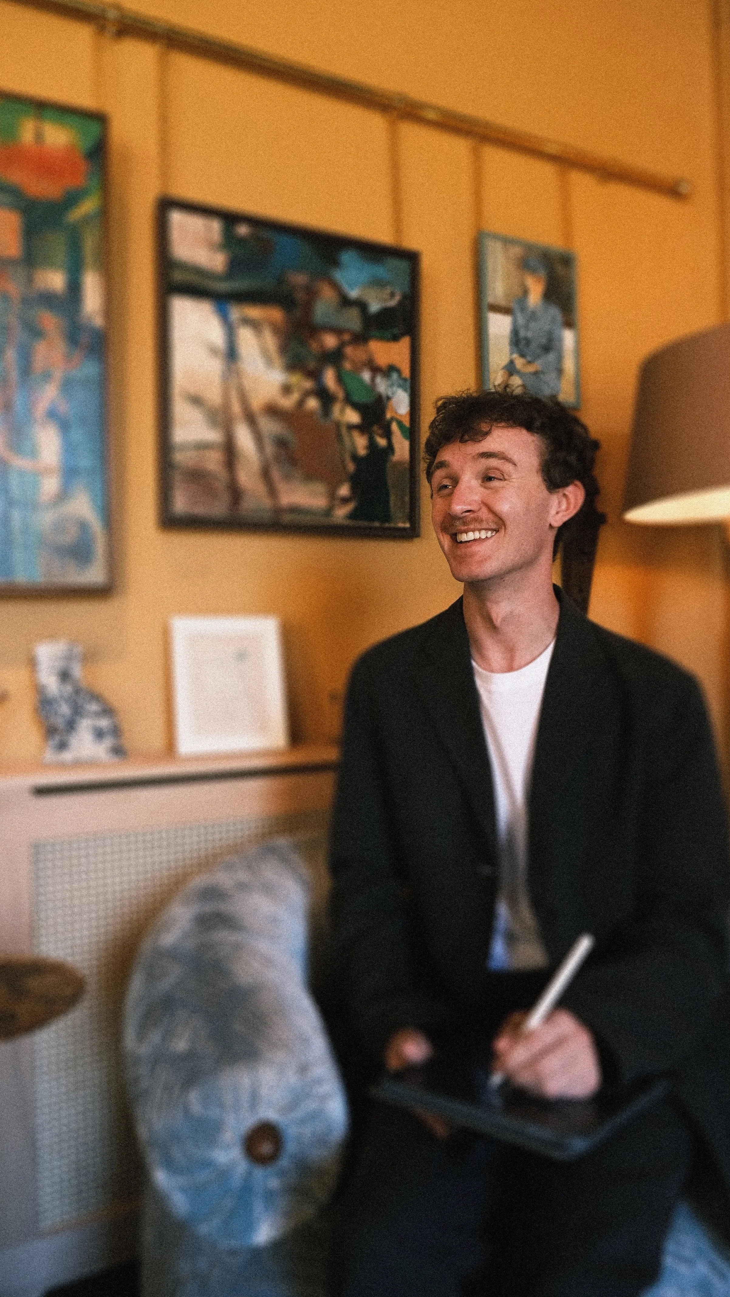 A smiling man with curly hair sitting on a couch holding a digital stylus and a tablet, with framed artwork on a yellow wall behind him.
