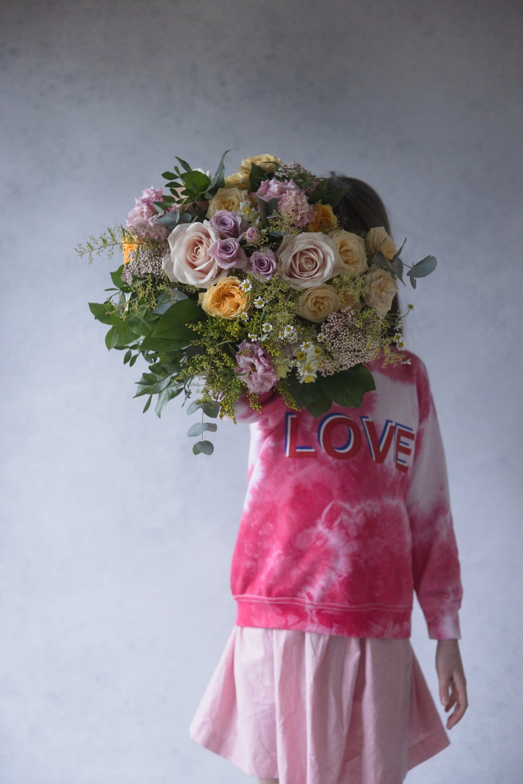 Colorful bouquet of various fresh flowers including roses, carnations, and lilies arranged on a white marble-topped stand with a pastel, floral, and curtain background.