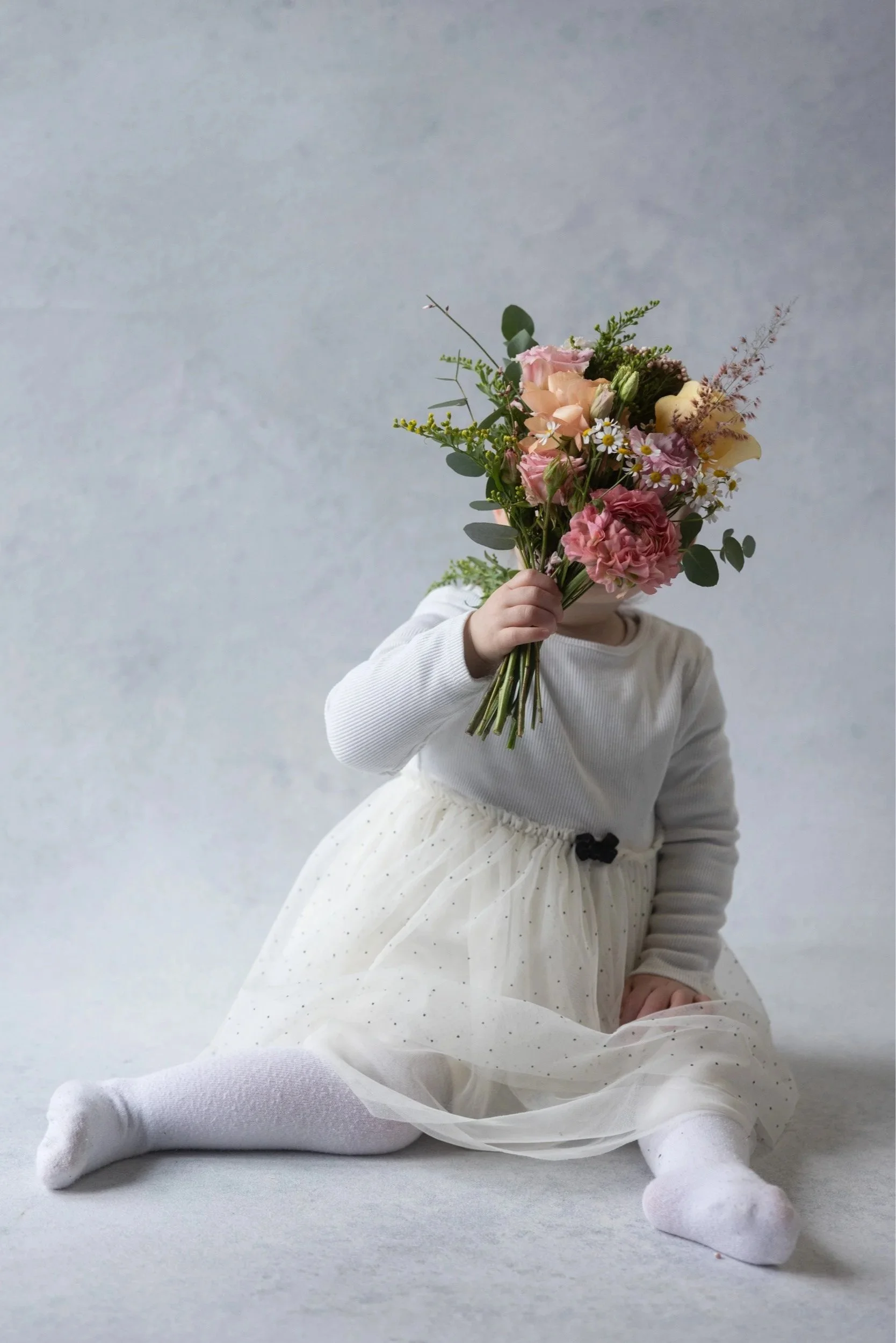 Child with blonde hair and red pajamas sitting on a black surface, surrounded by flowers shaped like a heart, with a paper letter 'I' and the word 'Mum' written in chalk, a small stuffed animal, and a red toy cart filled with flowers.