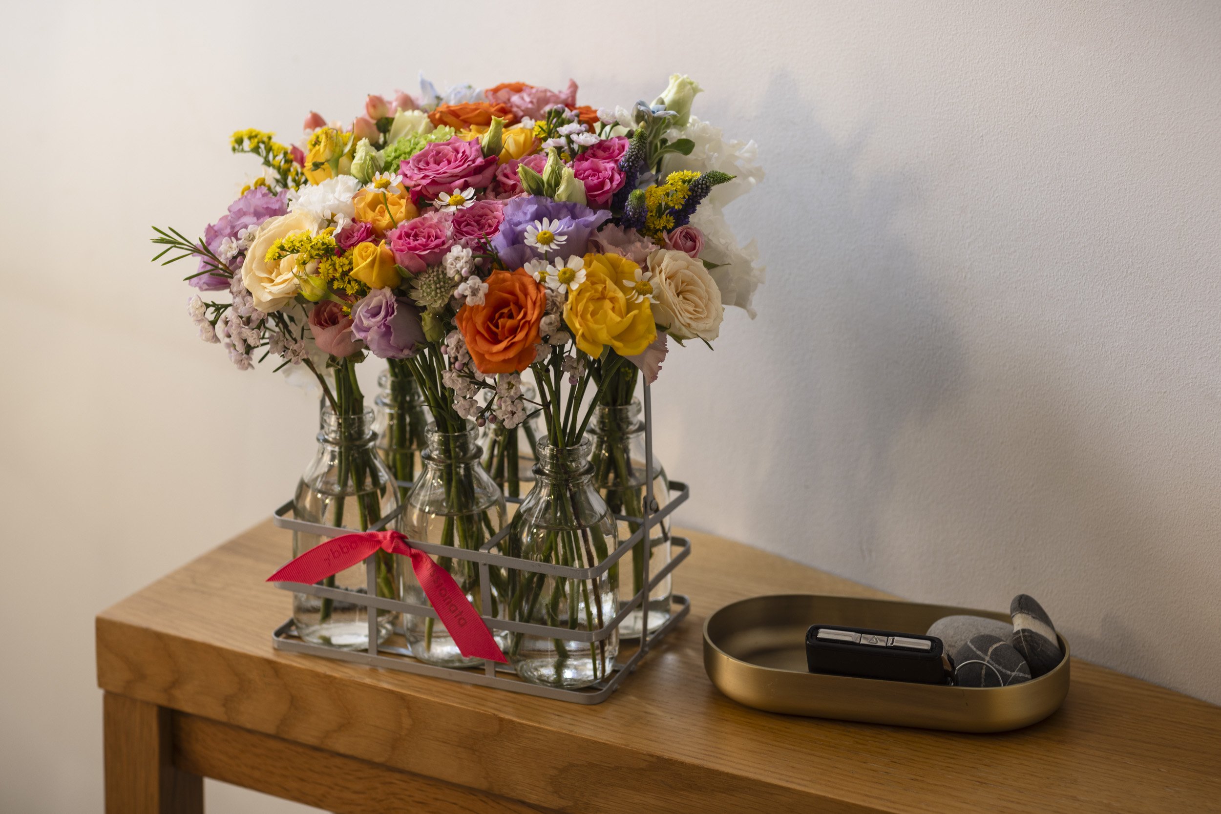 Person holding a metal basket with glass jars filled with pink, white, and cream-colored flowers, decorated with a pink ribbon.
