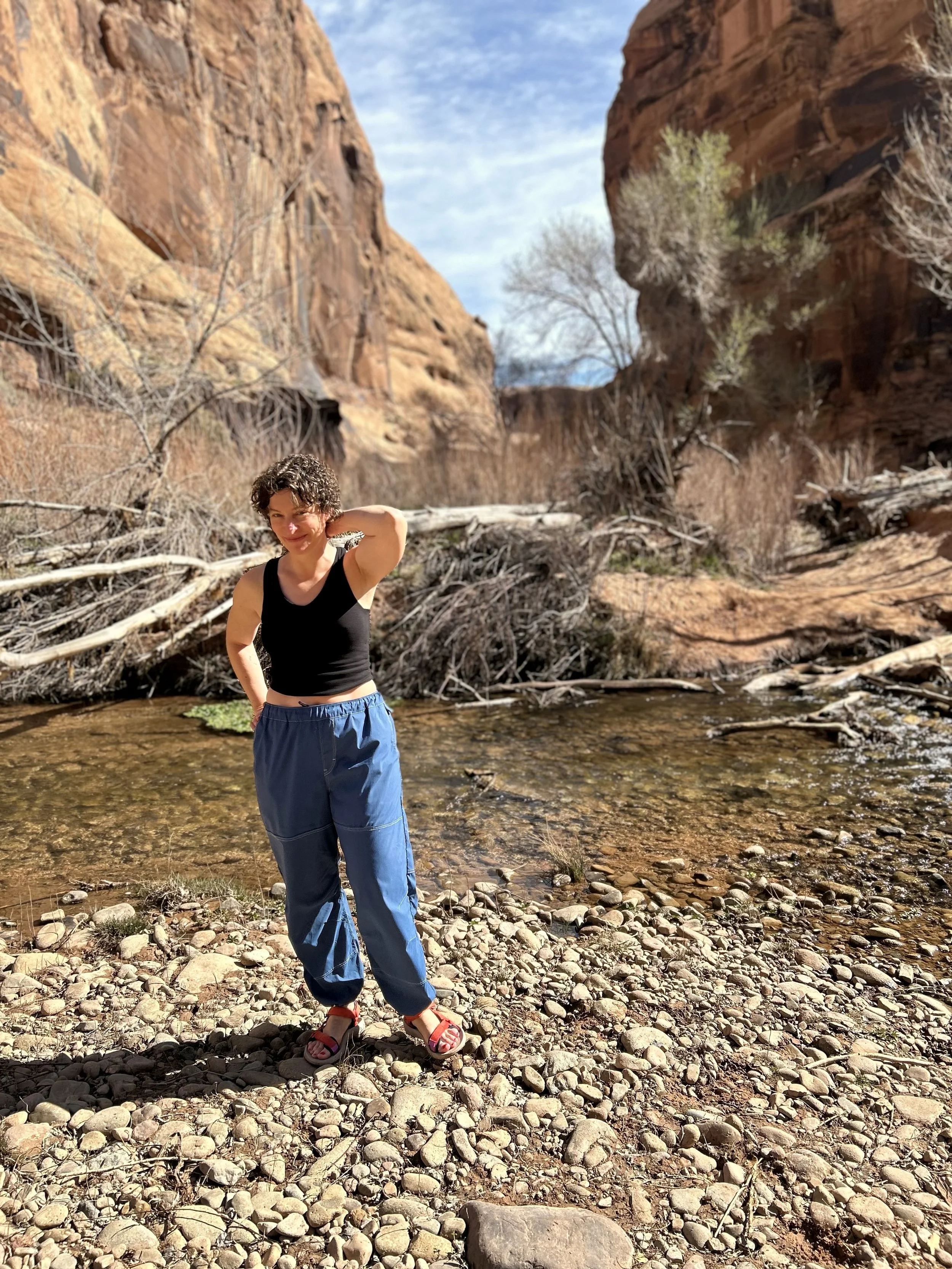A straight sized white woman poses on a riverbank by red cliffs.