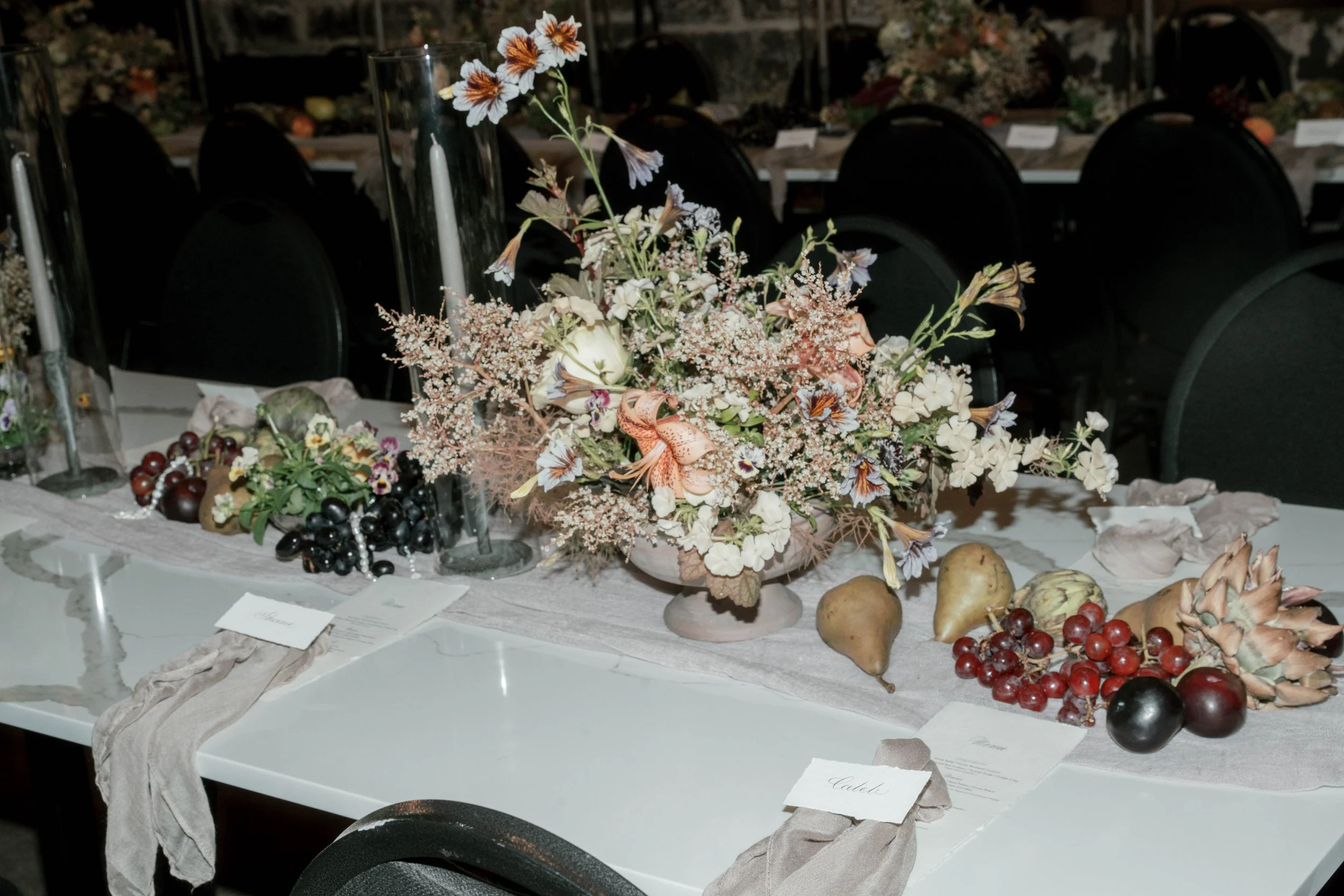A table centerpiece consisting of a large floral arrangement with light pink, white, and orange flowers, set on a table decorated with fabric runners, candles in glass holders, and placed with grapes, pears, artichokes, and place cards.
