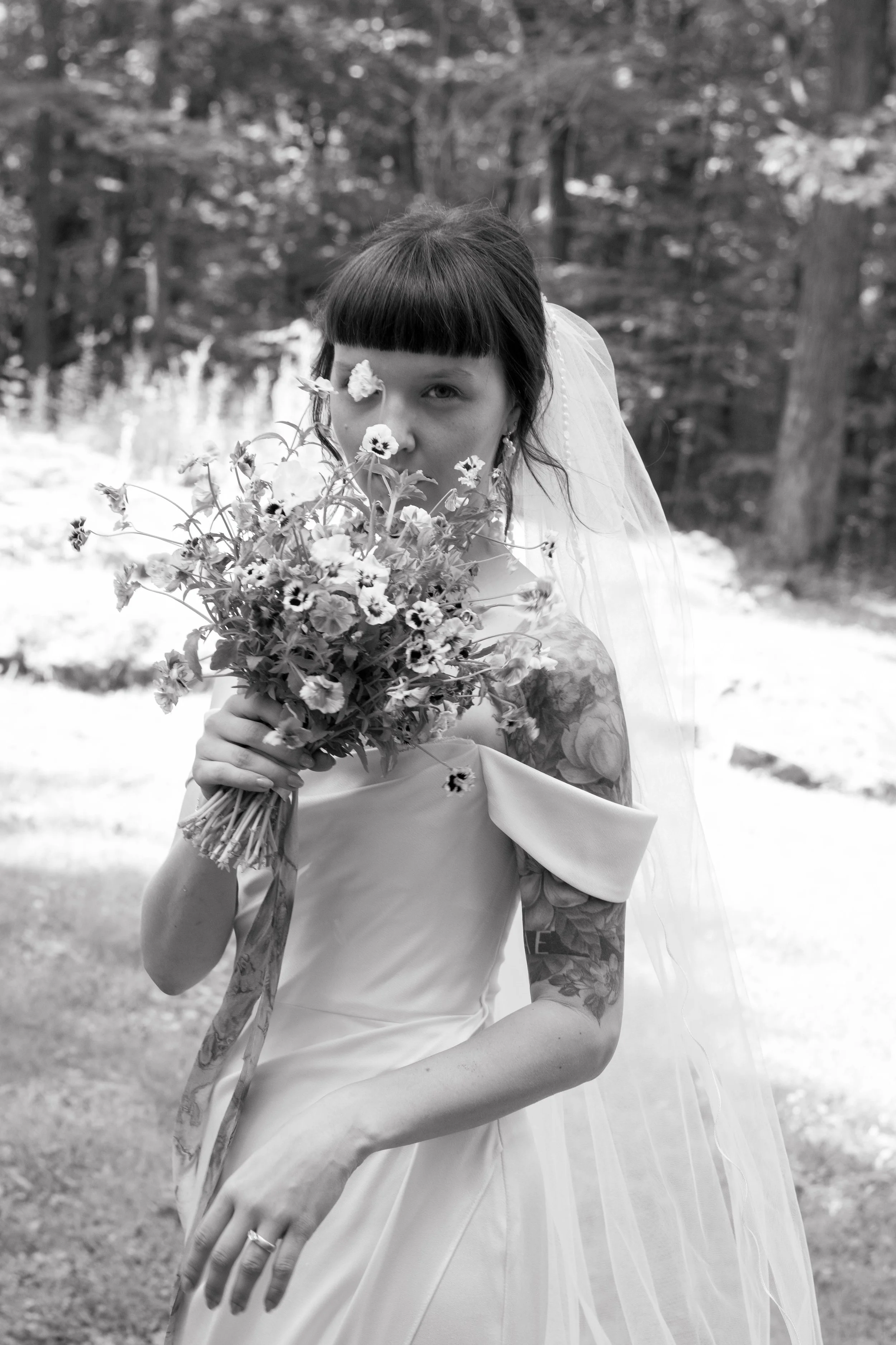 A woman with tattoos in a wedding dress holding a large bouquet of flowers, standing outdoors in a natural setting.