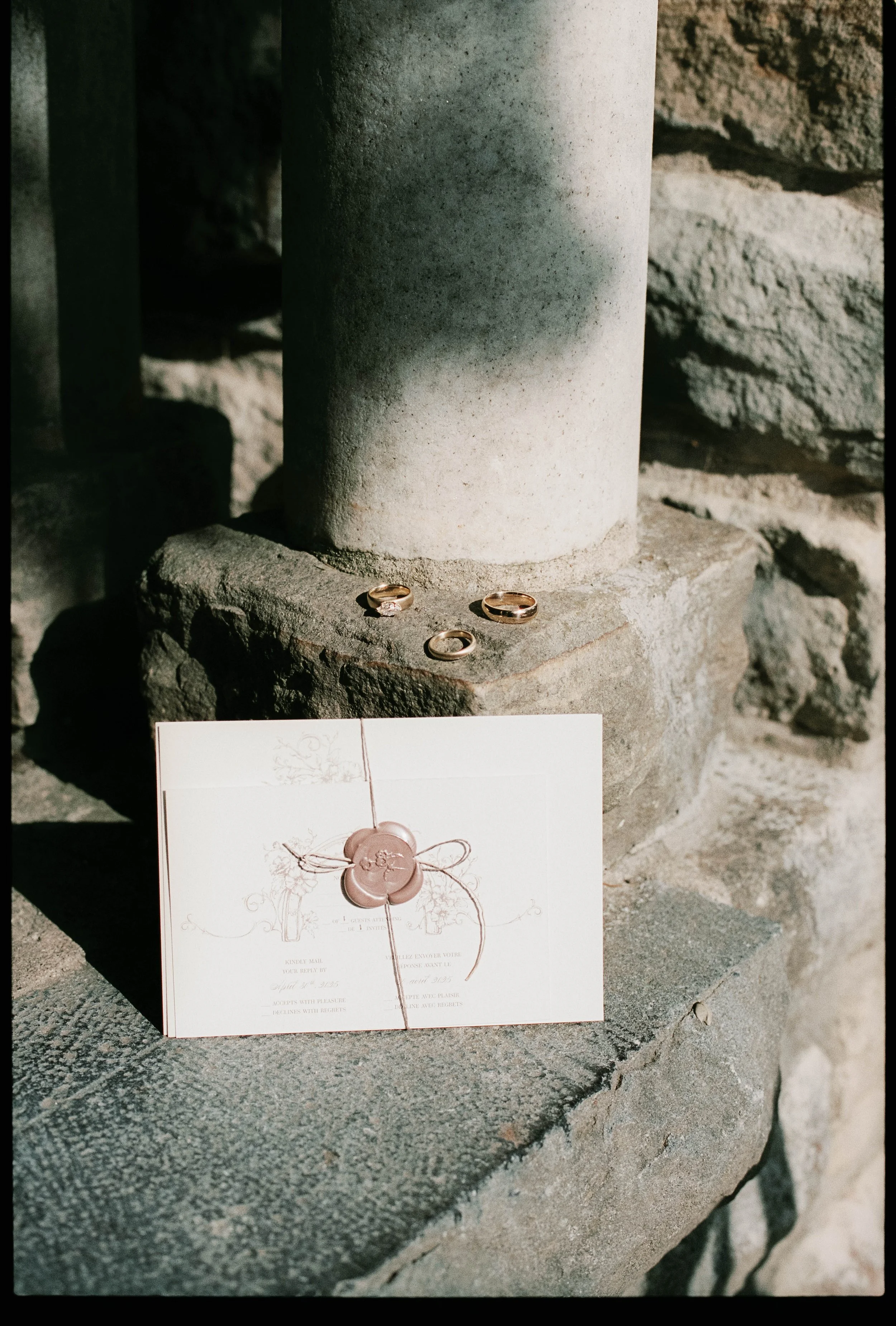 Three wedding rings and a wedding invitation with a wax seal placed on a stone ledge in front of a stone pillar.