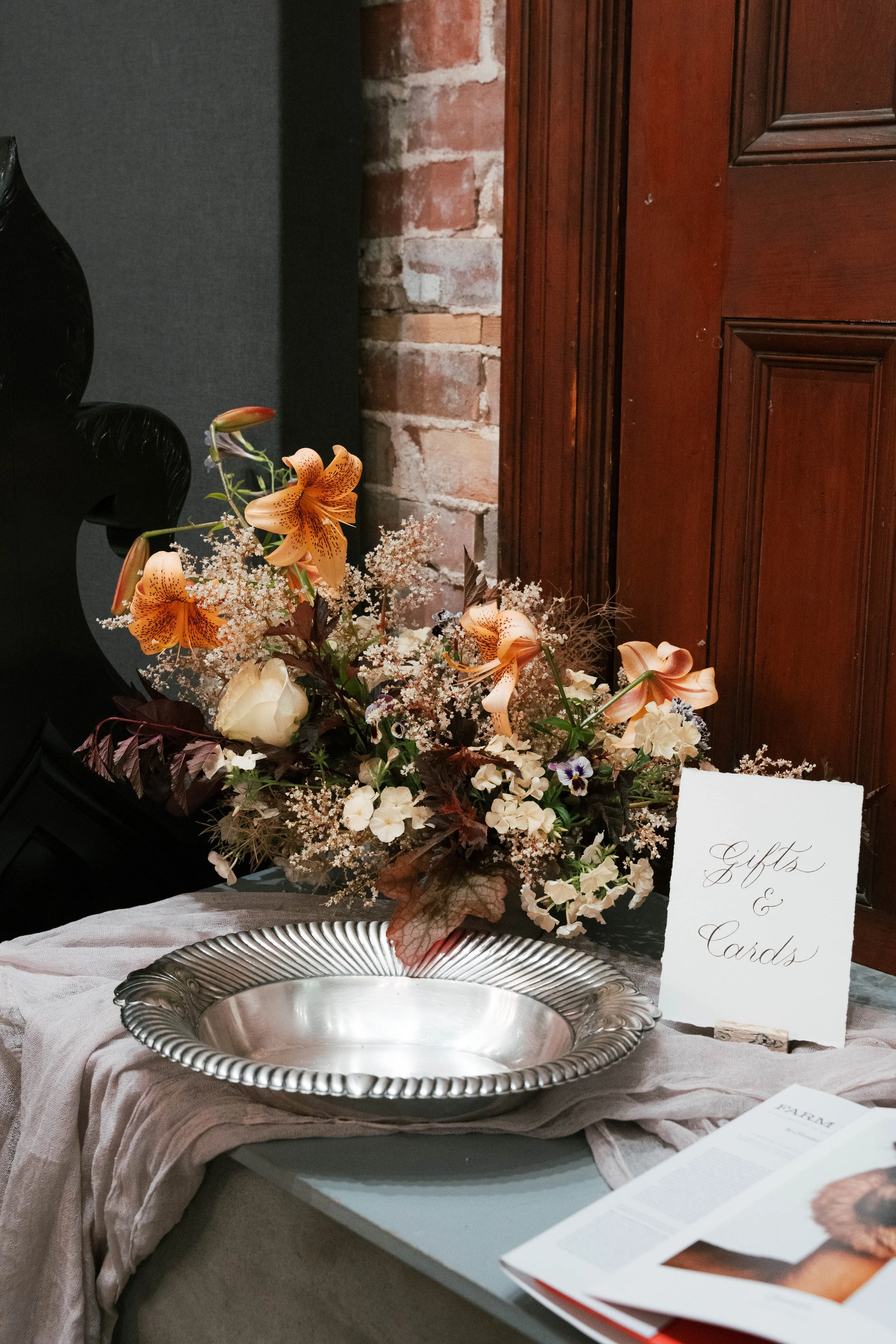 A floral arrangement with lilies, roses, and other flowers on a table with a sign that says 'Gifts & Cards'.
