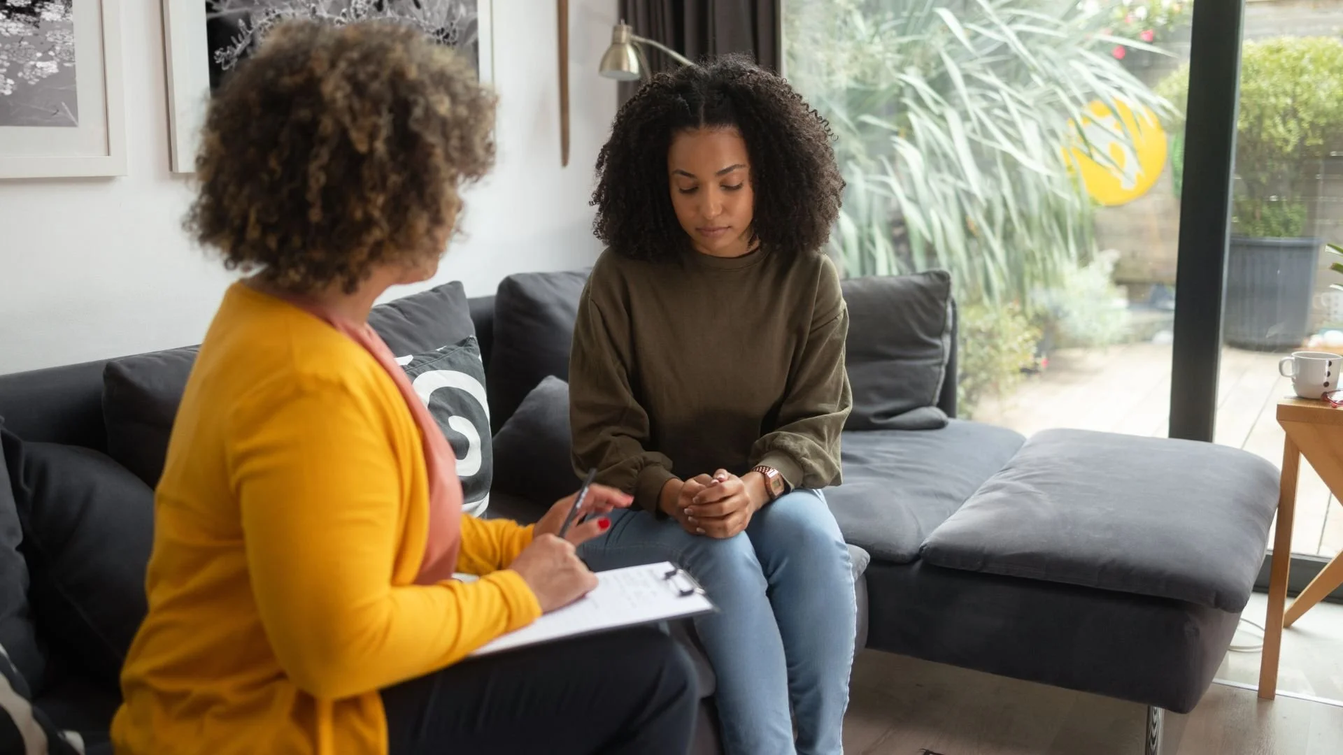 A young Black woman looking down thoughtfully during a mental health counseling session with a therapist who is holding a clipboard and taking notes in a bright office.