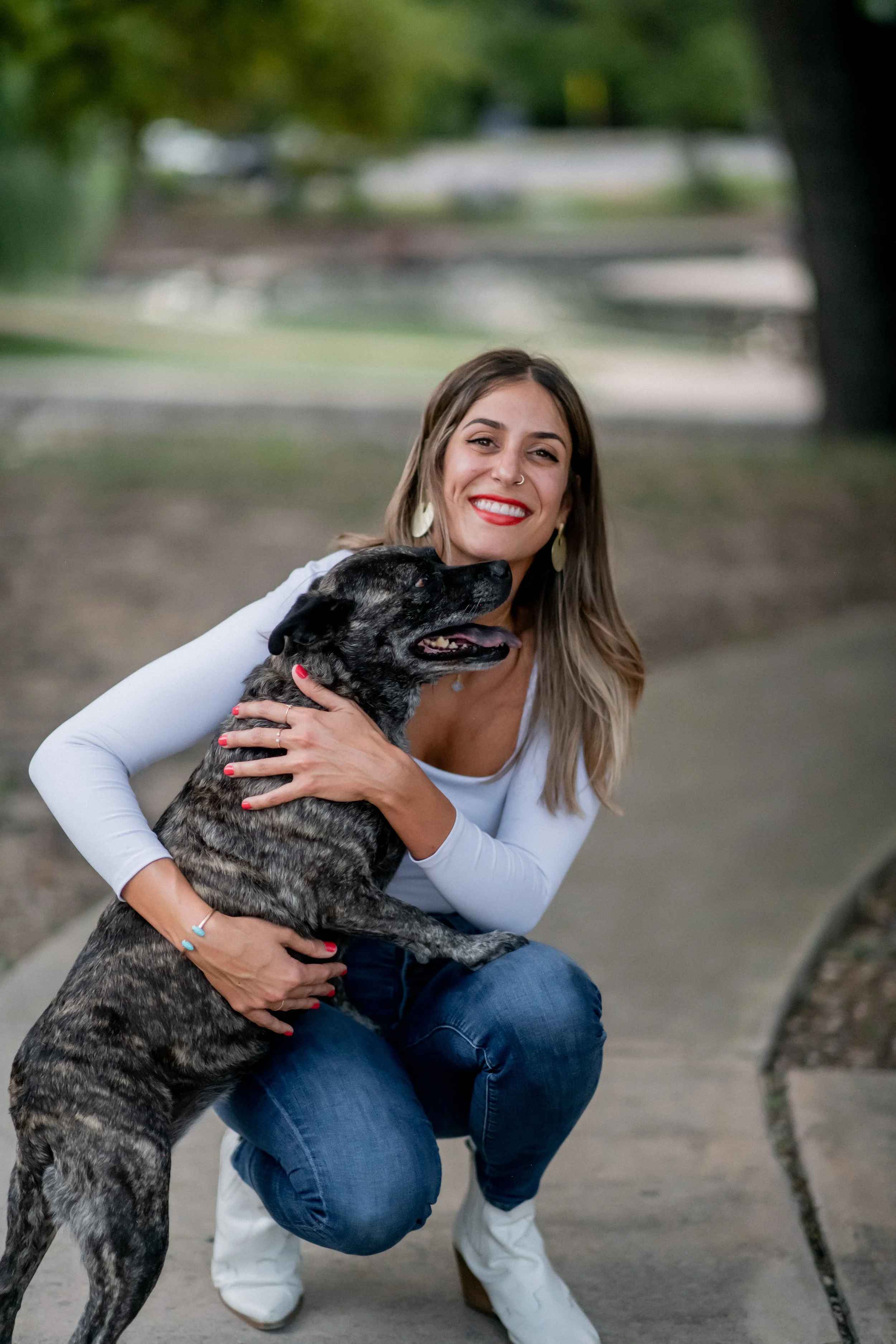 A woman with long, light brown hair, red lipstick, wearing a white long-sleeve shirt, blue jeans, white boots, and large earrings, is smiling and hugging a large brindle dog with pointed ears and a black nose in an outdoor park setting.