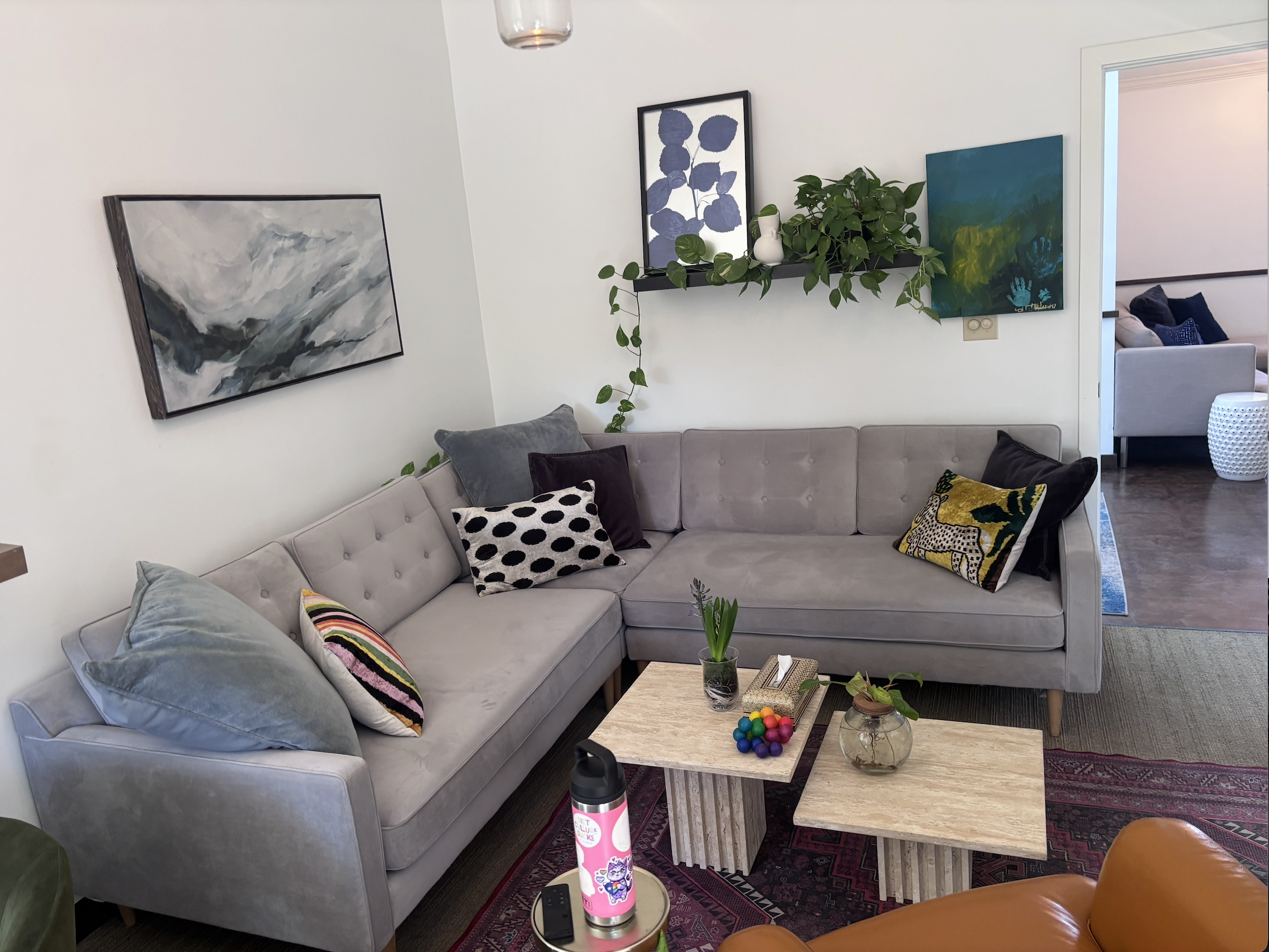Living room with gray sectional sofa decorated with various colorful pillows, a wooden coffee table with a glass vase and colorful balls, and art on the walls. There is a large painting of a mountain scene, a shelf with plants and vases, and a partial view of another room with seating and cushions.