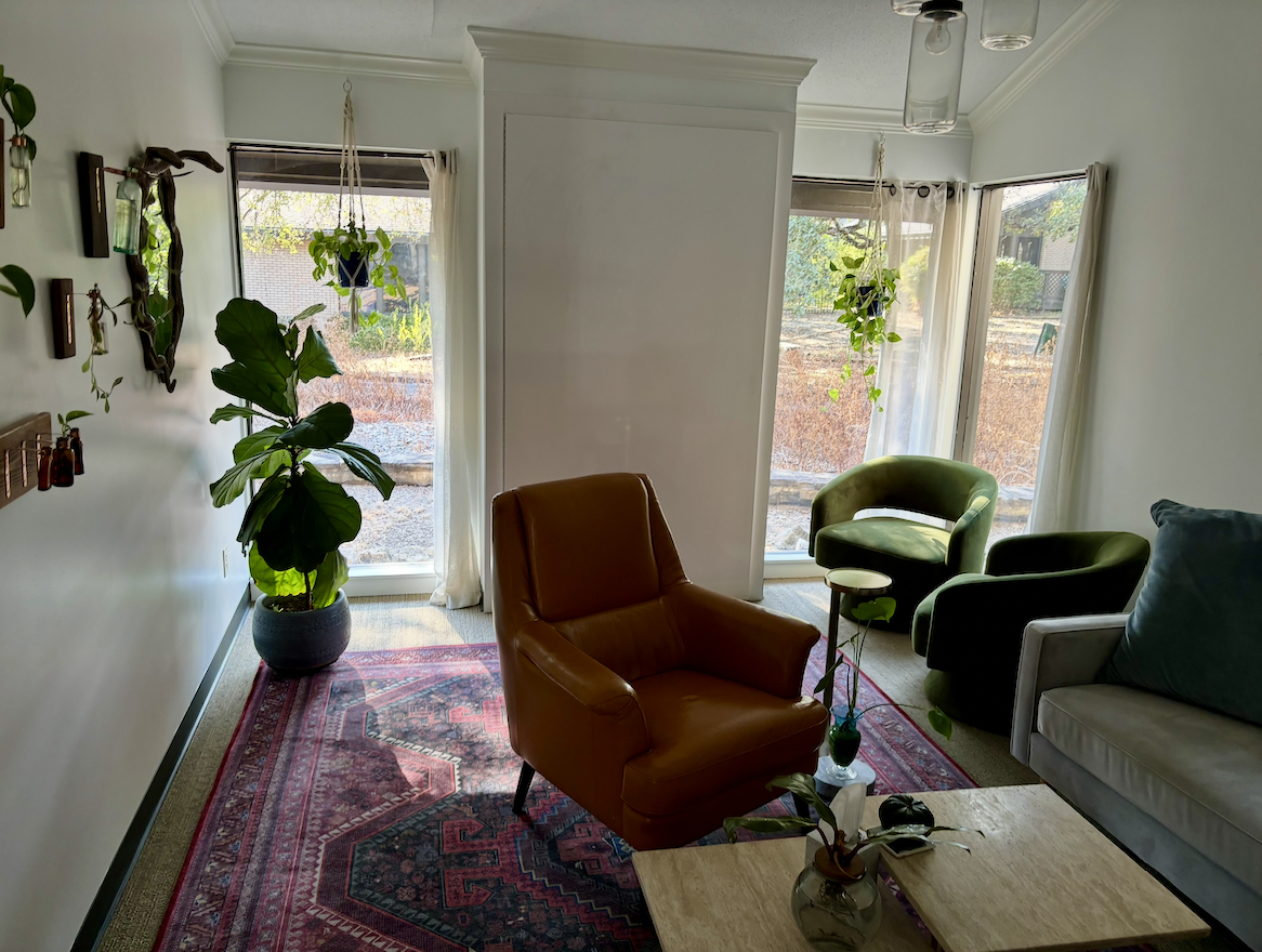Living room with large windows, a large potted plant, a red armchair, a green chair, a gray couch, a wooden coffee table with decorative plants, and hanging plants from the ceiling.
