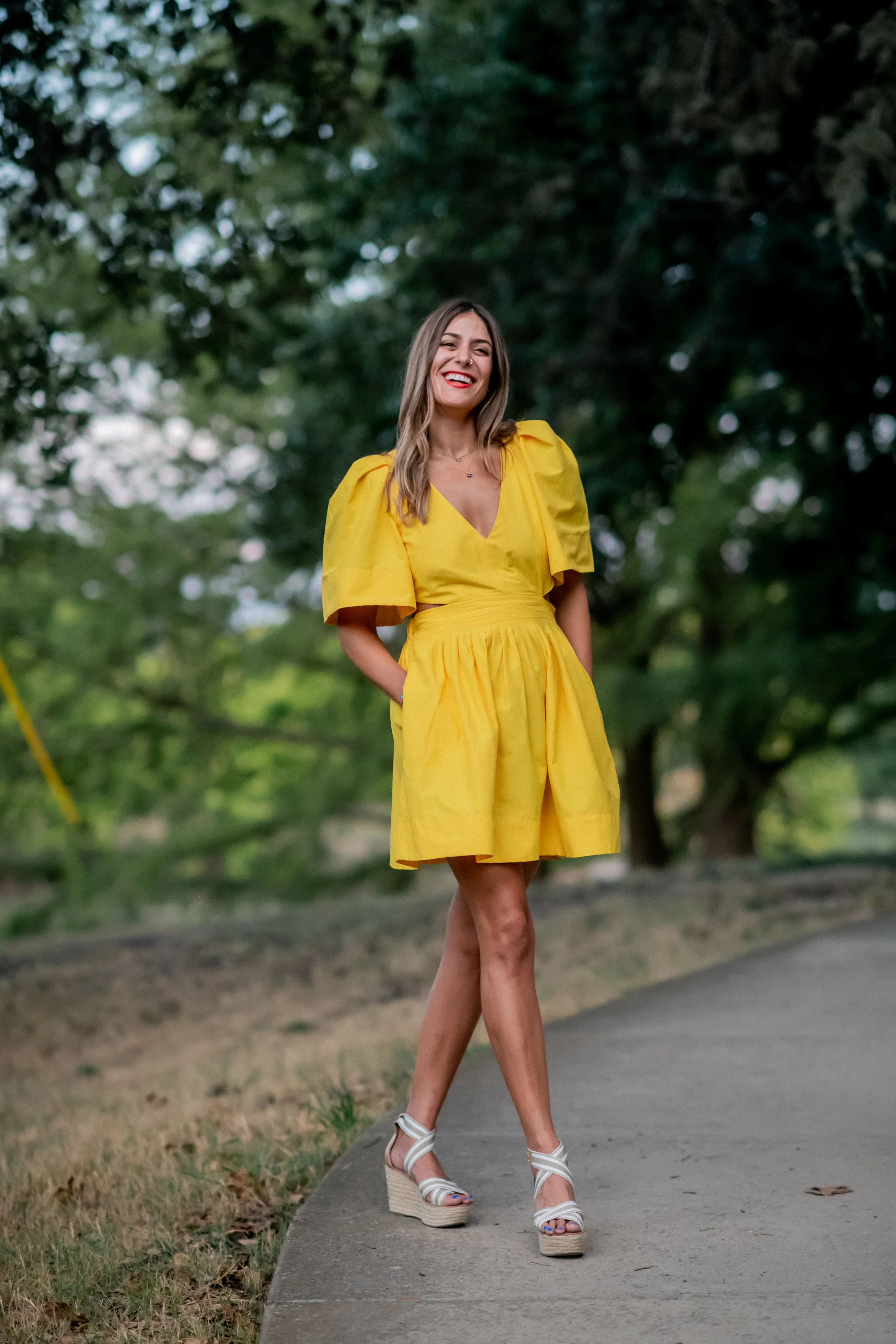 A woman in a yellow dress with puffed sleeves and a v-neckline walking on a sidewalk in a park, smiling with her hands behind her back, wearing white high-heeled sandals.