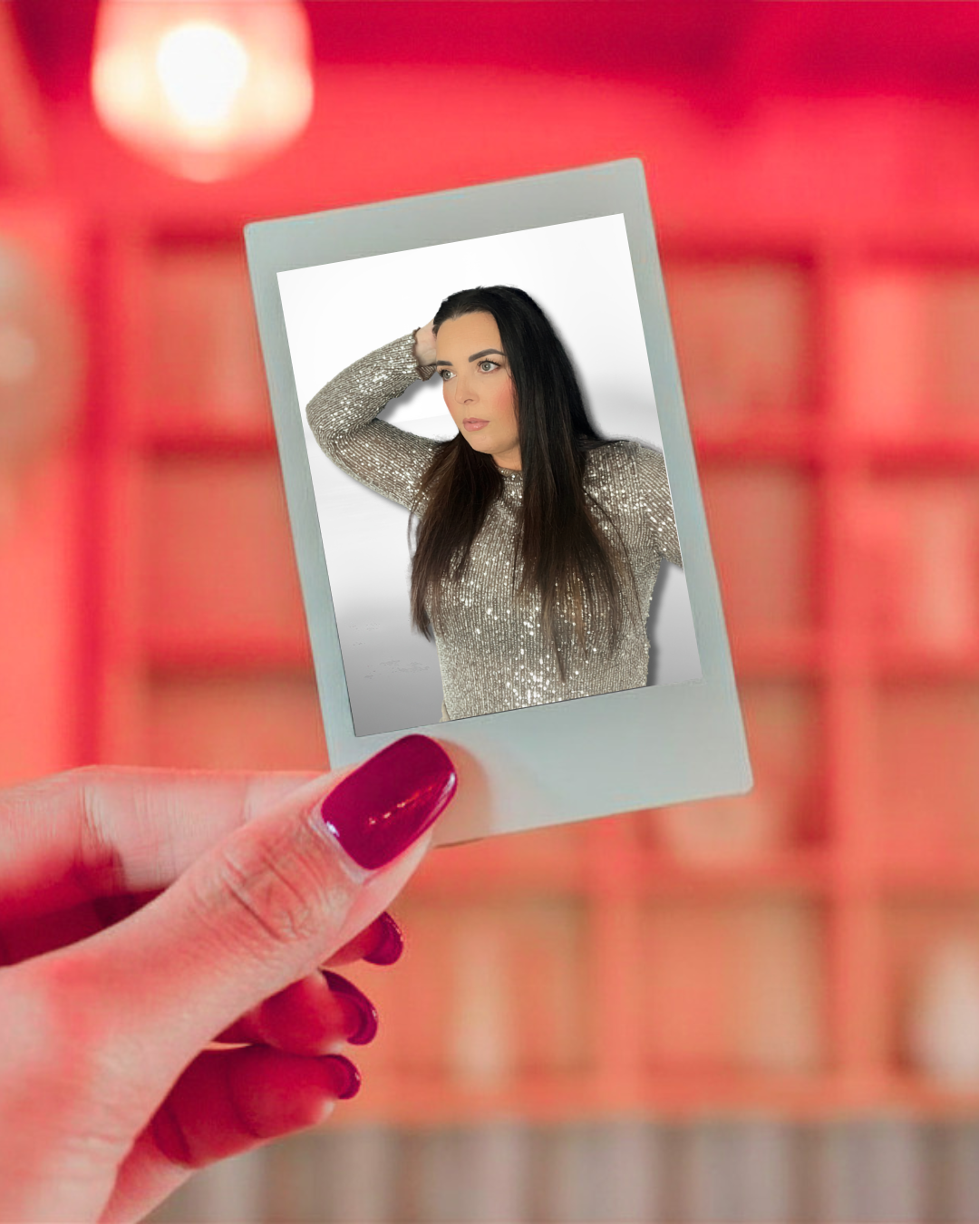 A hand with red nails holding a photo of a woman with long dark hair, wearing a sparkly top, against a blurred red background.