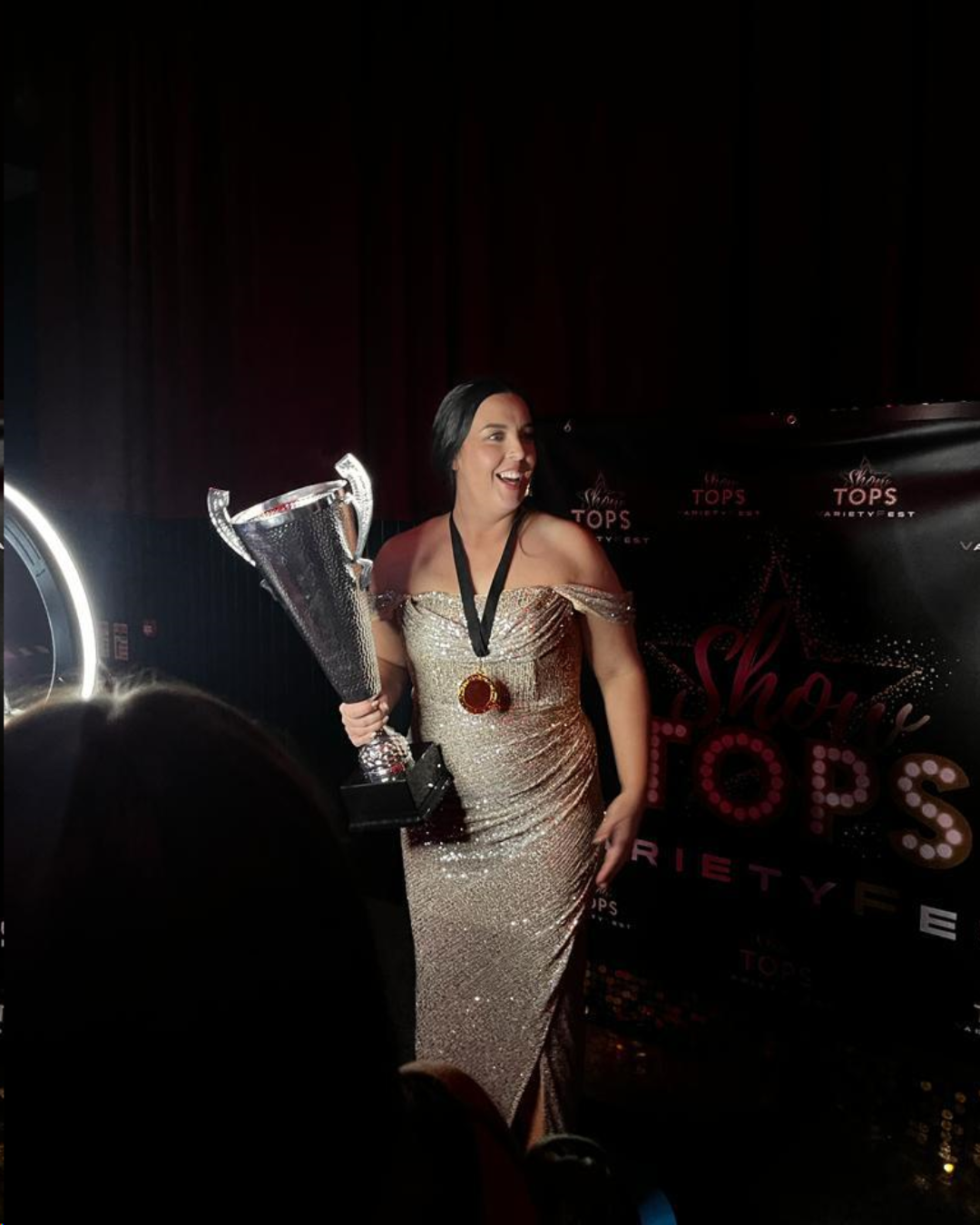 Woman in a gold sequined gown holding a large silver trophy, standing in front of a black backdrop with event logos, wearing a medal around her neck, smiling.