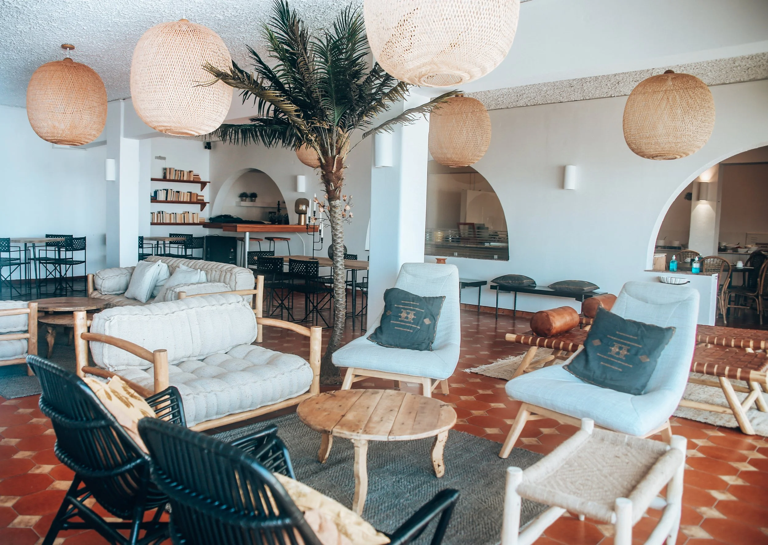 Sala de estar con decoración bohemia, sofás y sillas de mimbre, mesa de centro de madera, alfombra y lámparas colgantes, con estante de libros en el fondo.