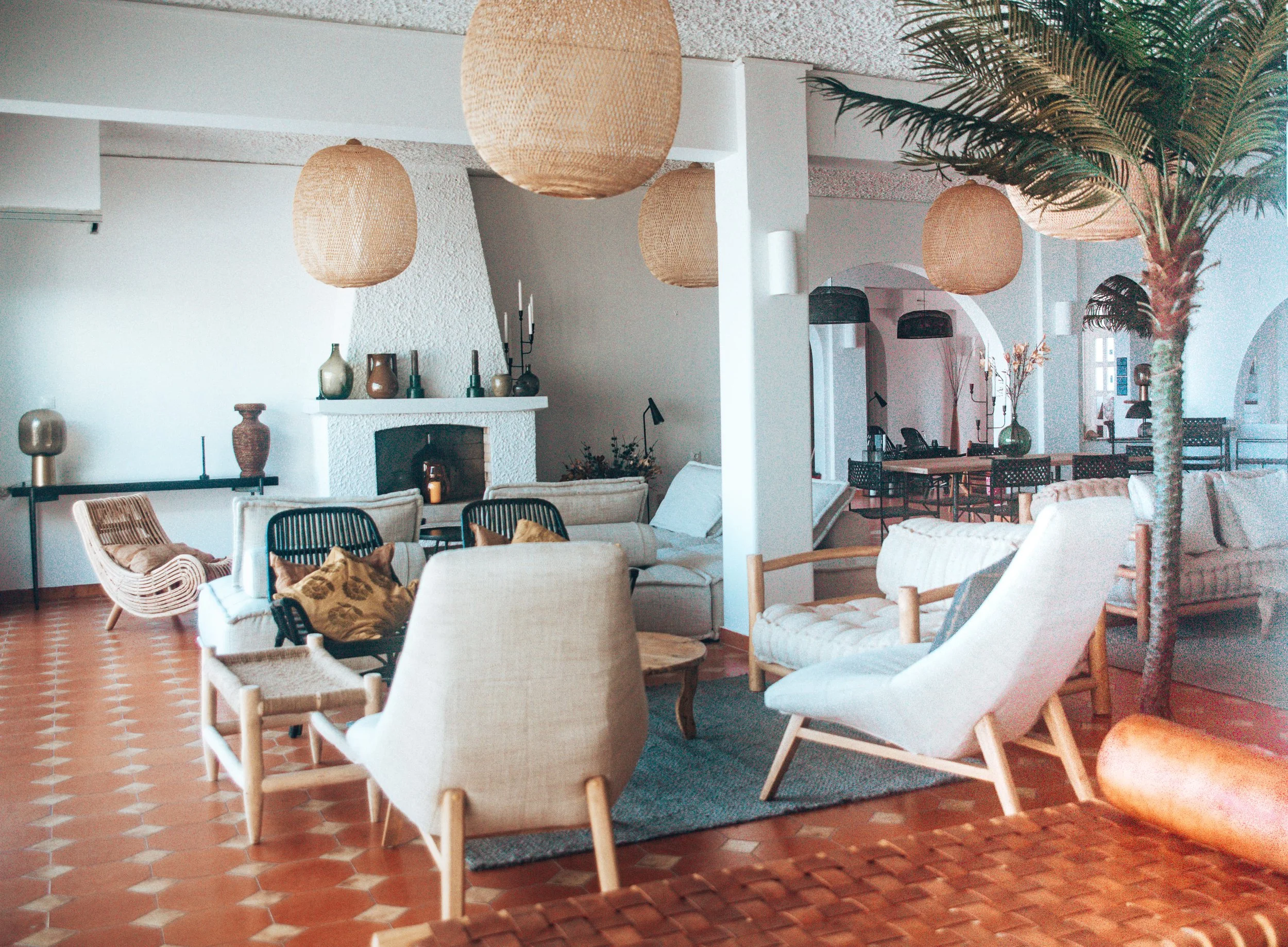 Sala de estar con decoración moderna, incluye muebles de madera y ratán, lámparas colgantes, chimenea, plantas y piso de baldosas naranjas.