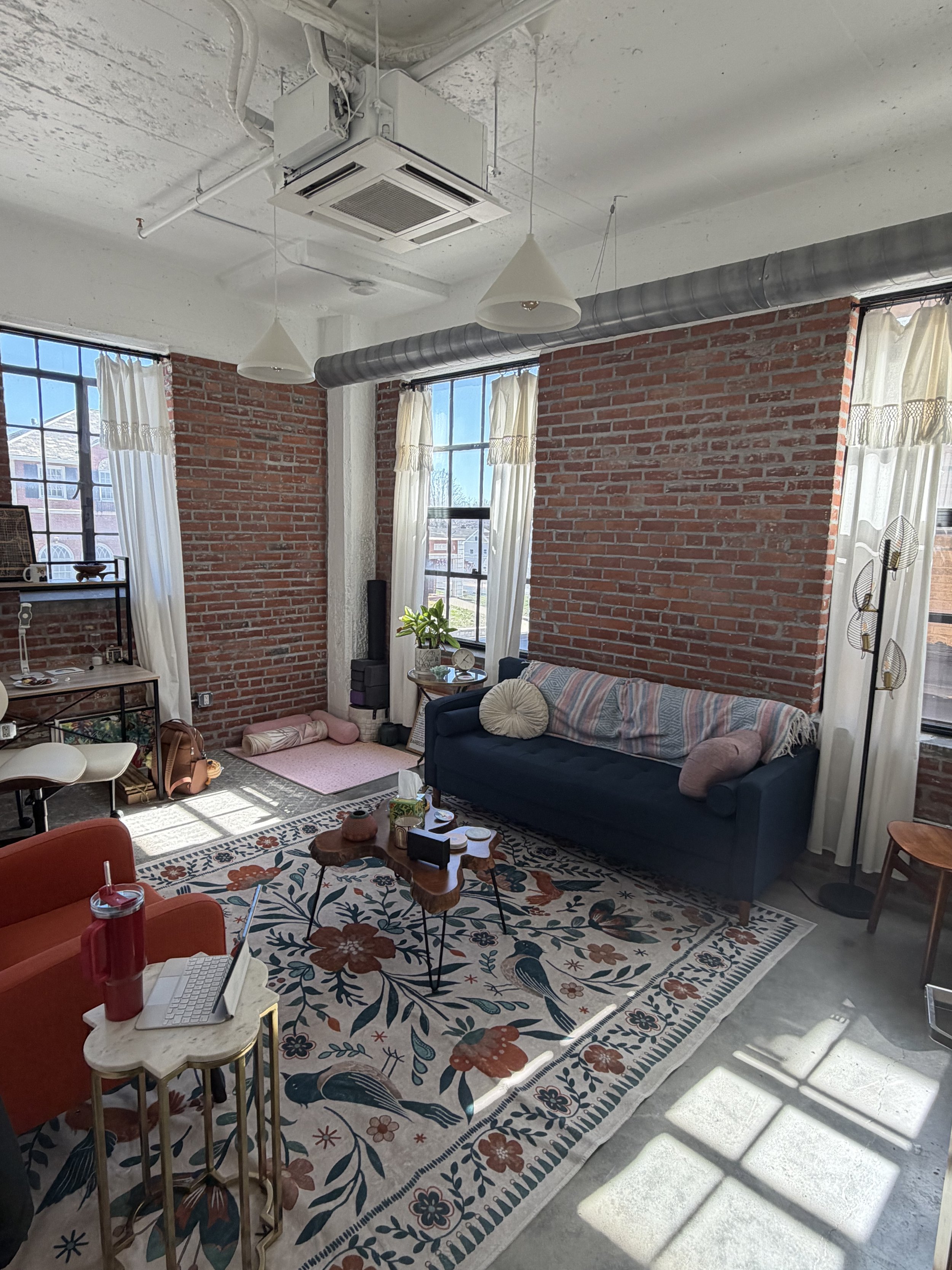 Bright living room with exposed brick walls, large windows with white curtains, and eclectic furniture including a navy sofa with patterned pillows, an orange armchair, and a small wooden coffee table with candles and decor. Sunlight streams through the windows onto a floral area rug.