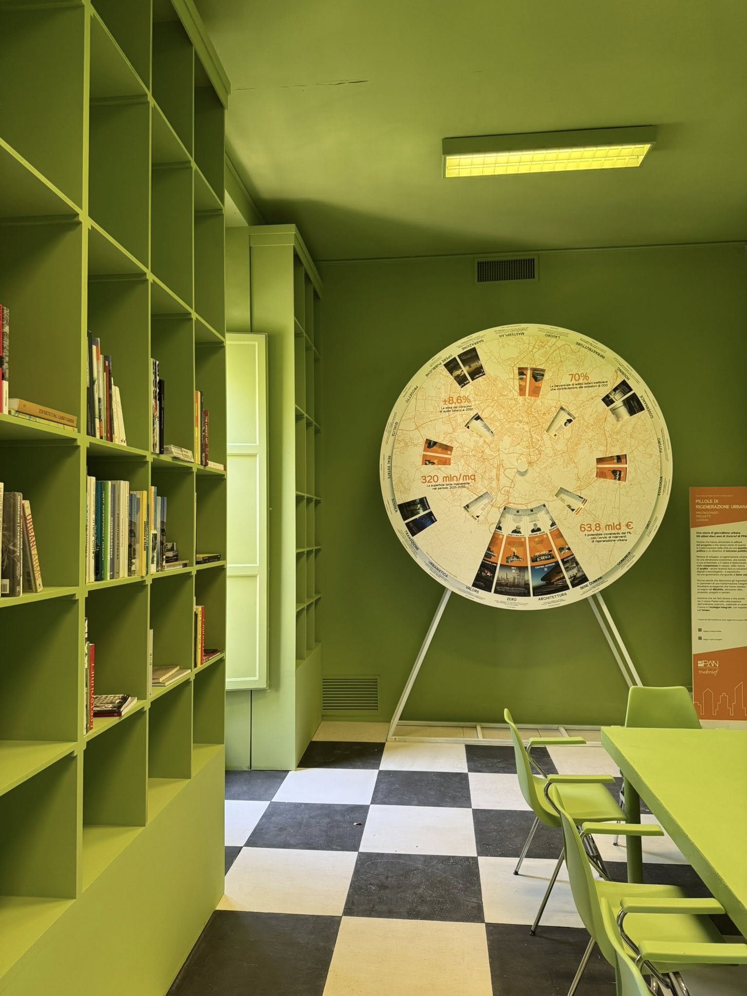 Reading area of the Countless City Library, with green shelving, tables and a circular visual archive on the wall.