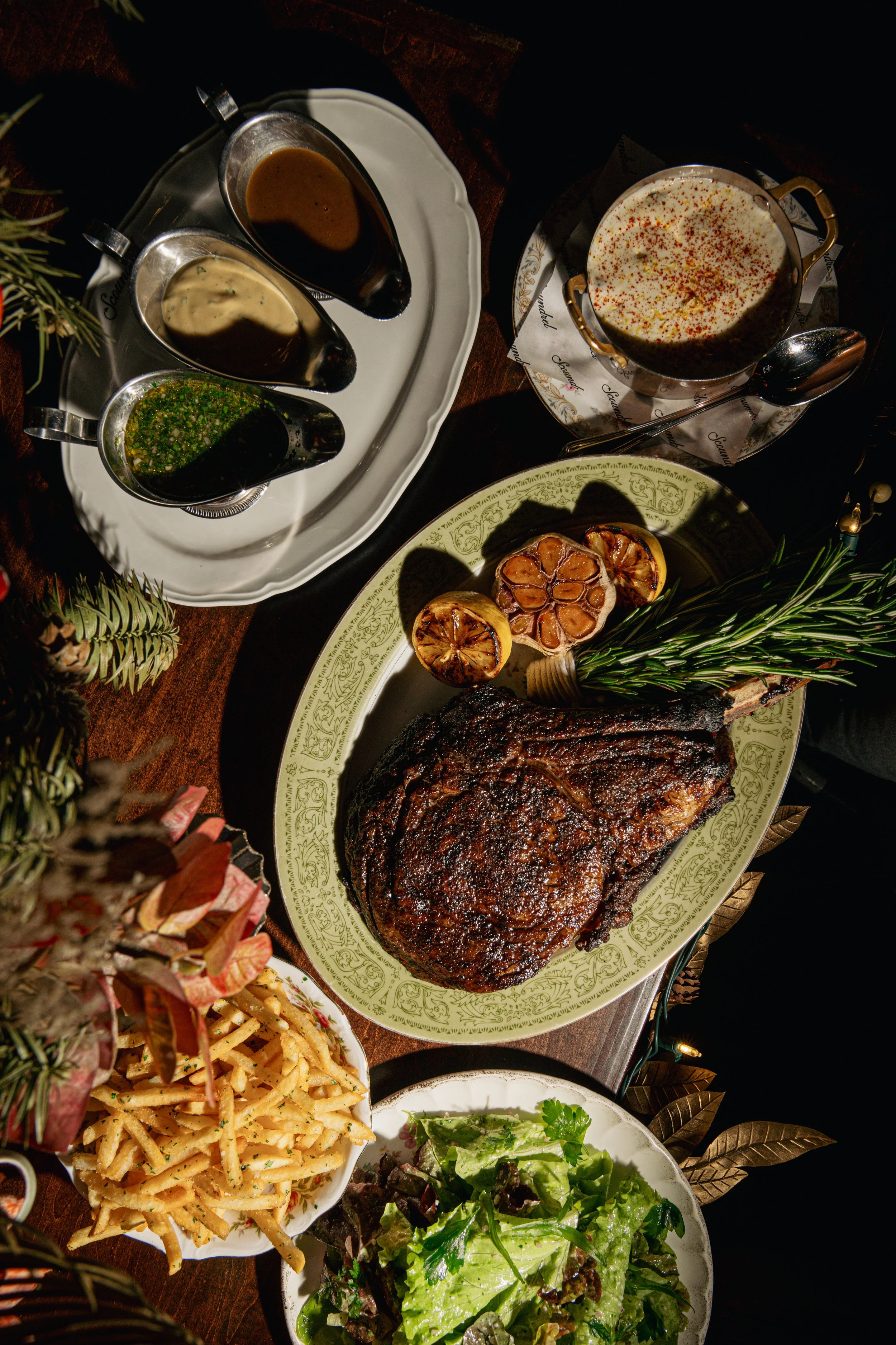 A roasted beef tomahawk on a platter garnished with garlic, lemon, and rosemary, accompanied by a green salad, french fries, various sauces.