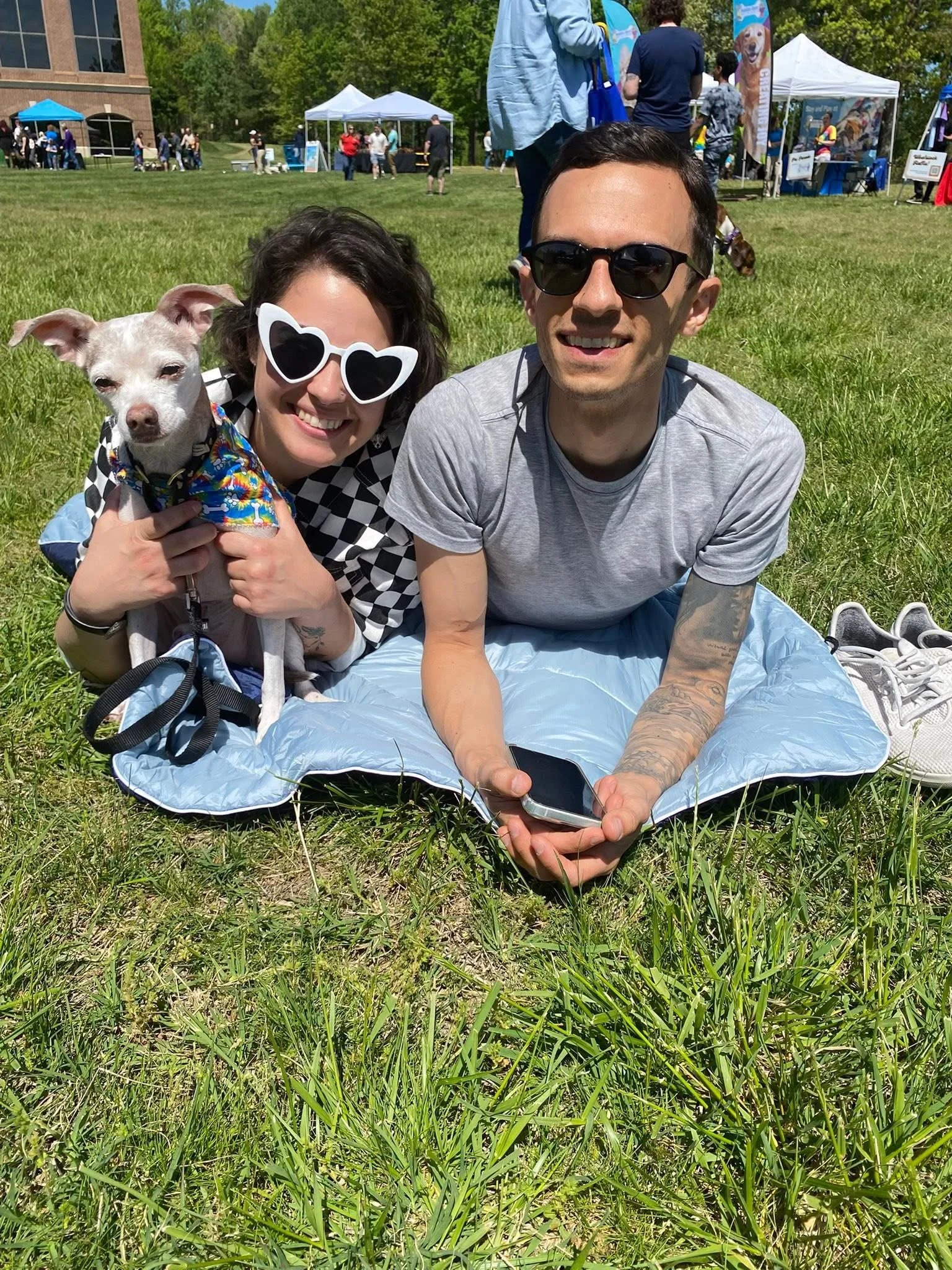 A smiling woman wearing sunglasses holding a small dog, and a man with dark hair and sunglasses lying on a blanket in a park at an outdoor event with tents and people in the background.