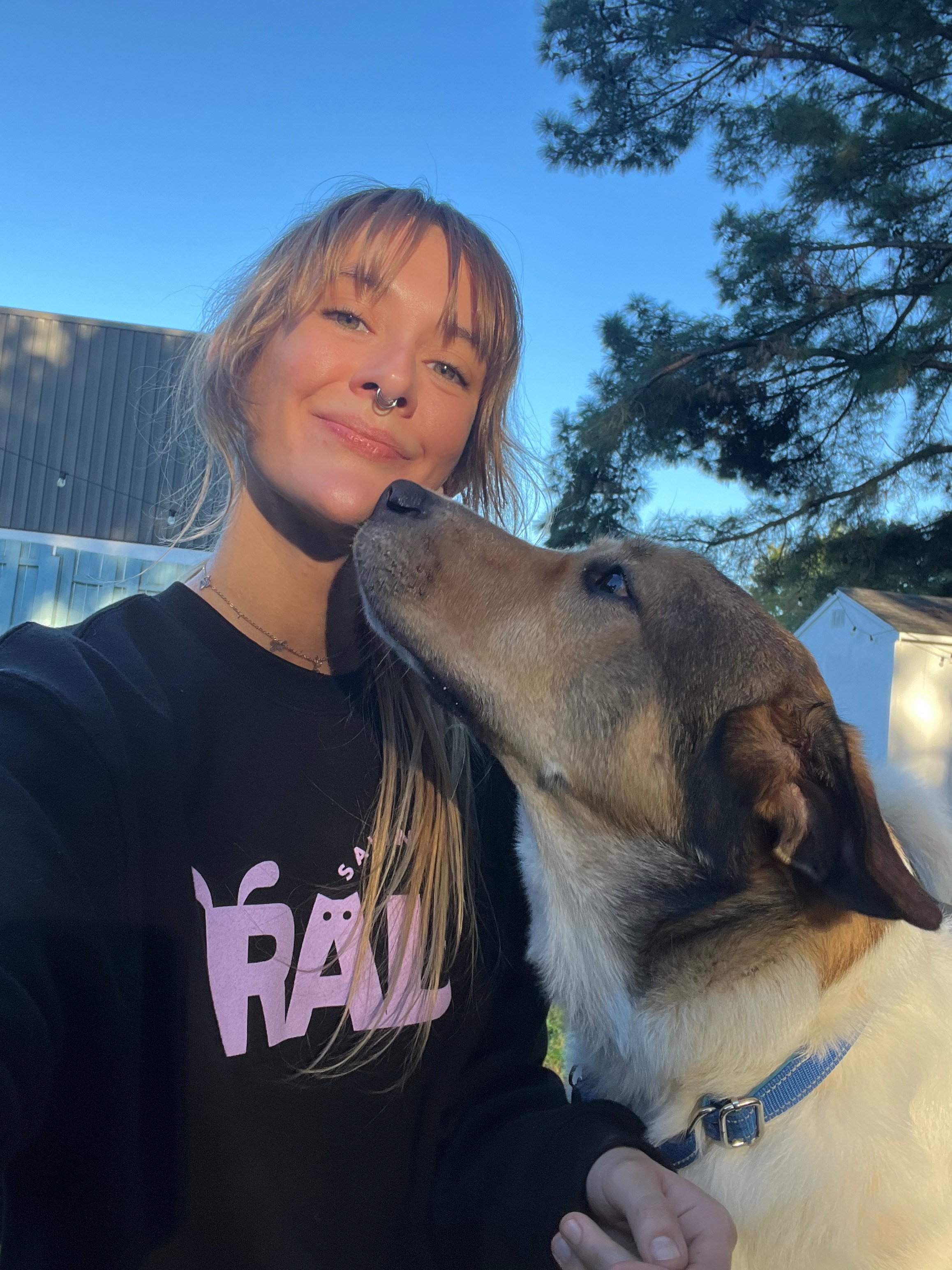 A woman with light brown hair and a septum piercing smiling as her dog sniffs her face outdoors during daytime.