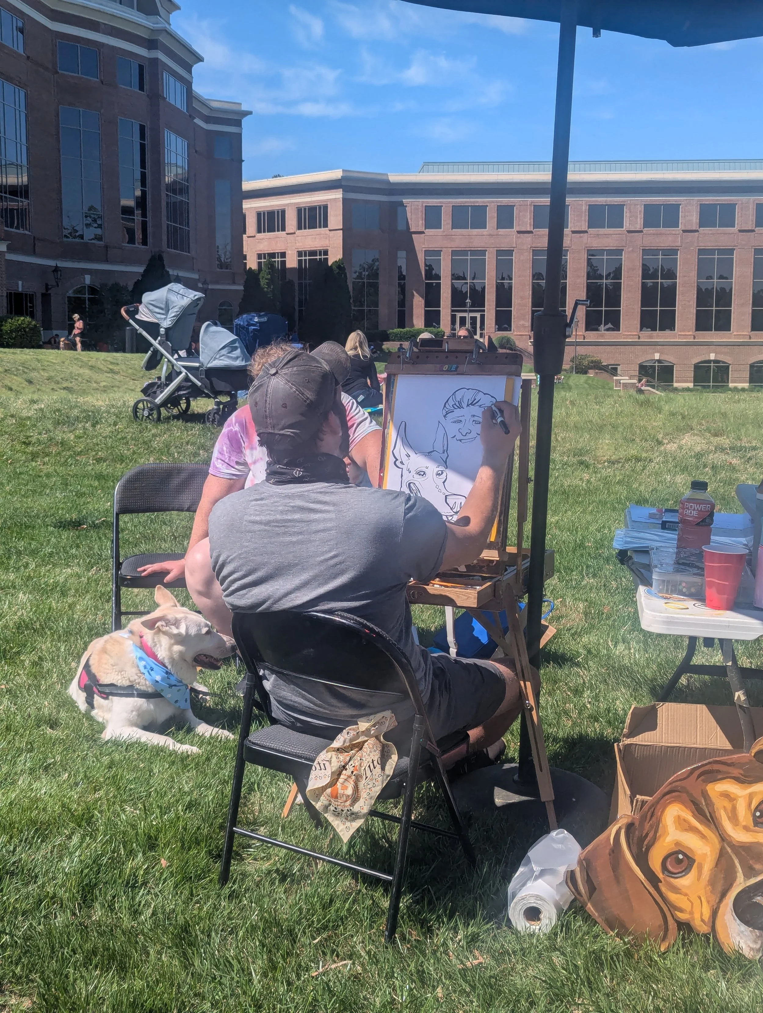 An artist drawing portraits of animals and people on an easel at an outdoor event on a grassy field, with a Labrador Retriever dog sitting nearby and a folding chair, a table with supplies, and a backpack in the scene.
