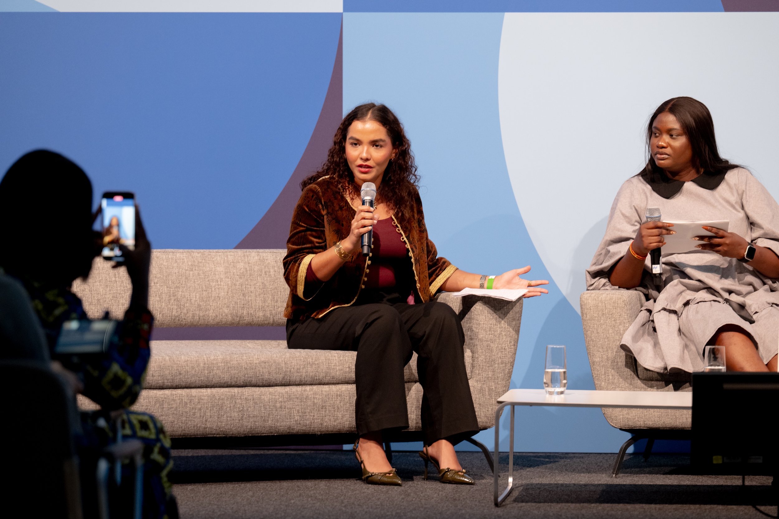 Two women are seated on a stage, participating in a panel discussion. The woman on the left is speaking into a microphone, wearing a brown velvet blazer and black pants. The woman on the right is holding a microphone and notes, wearing a light patterned dress with black collar details. There is a small table with a glass of water in front of them, and a person in the foreground is recording the event with a camera. The background features a blue and light blue abstract design.