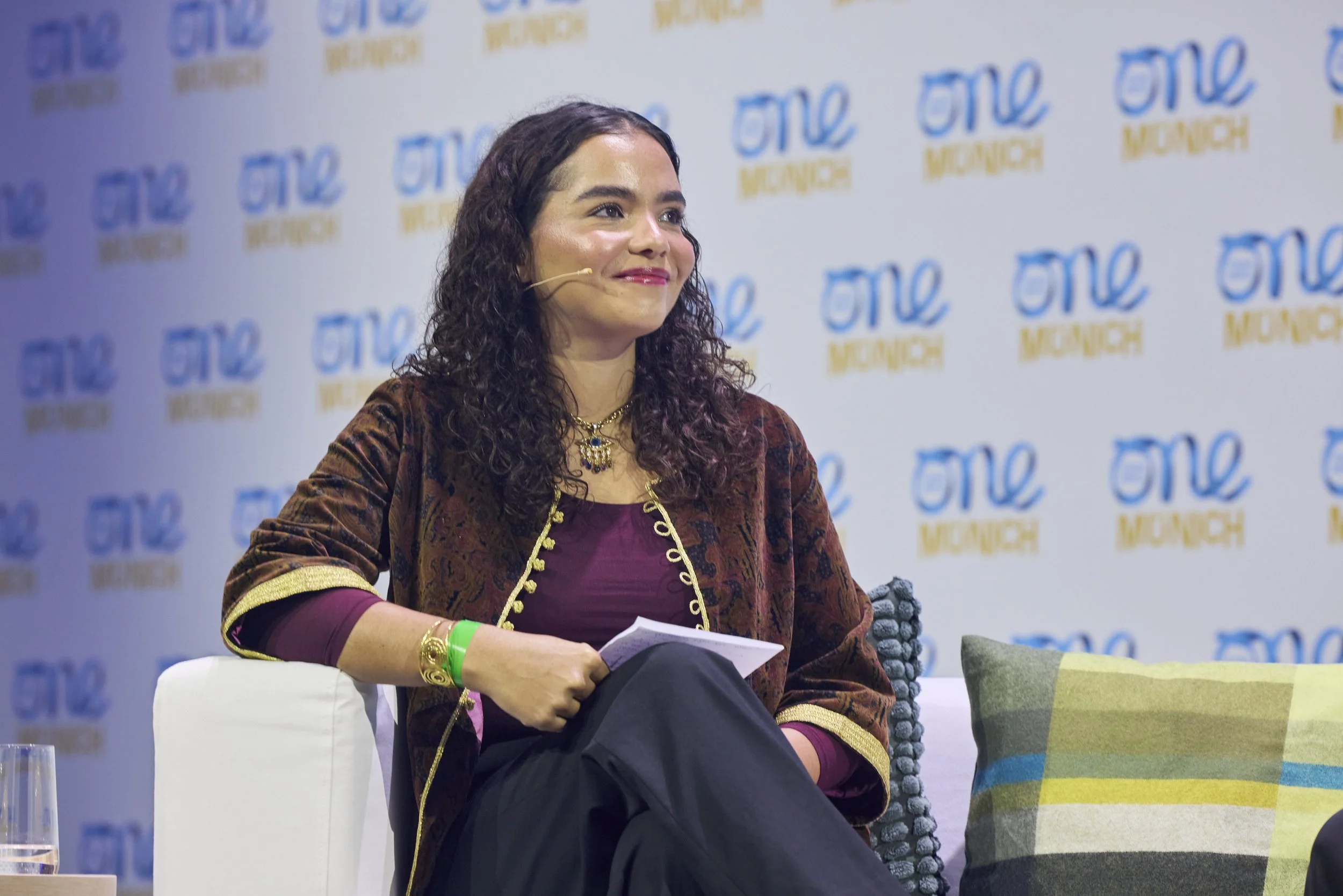 A woman with dark, curly hair smiling, wearing a traditional patterned jacket with gold details, seated on a stage at a panel or discussion event with a backdrop displaying the logo 'ONE WOMAN' repeatedly.