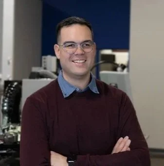 A man with glasses and a beard smiling indoors.