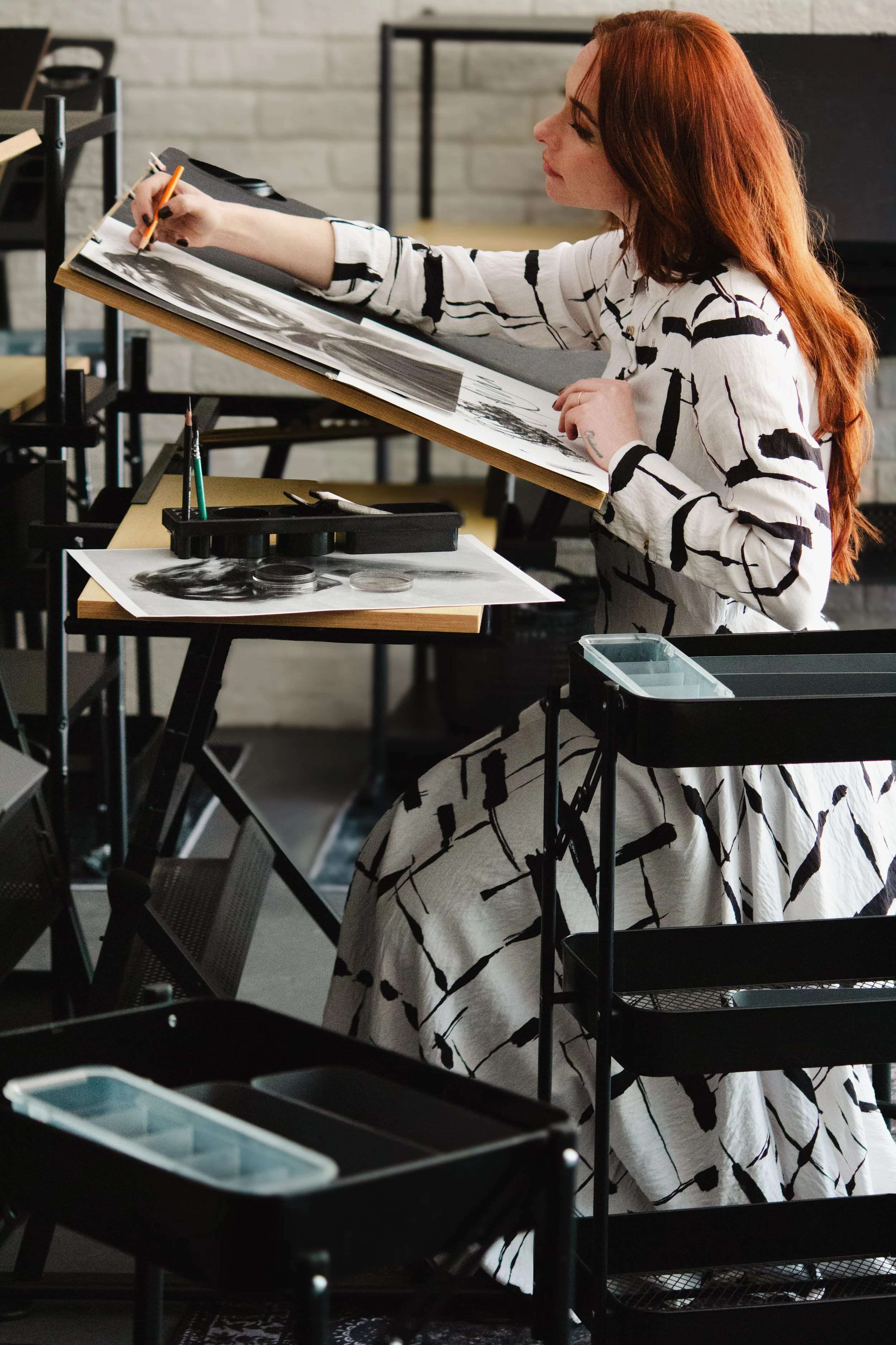 A person is drawing or working on a large  artwork at an art studio or classroom. There are art supplies and papers on the table and shelves around her.