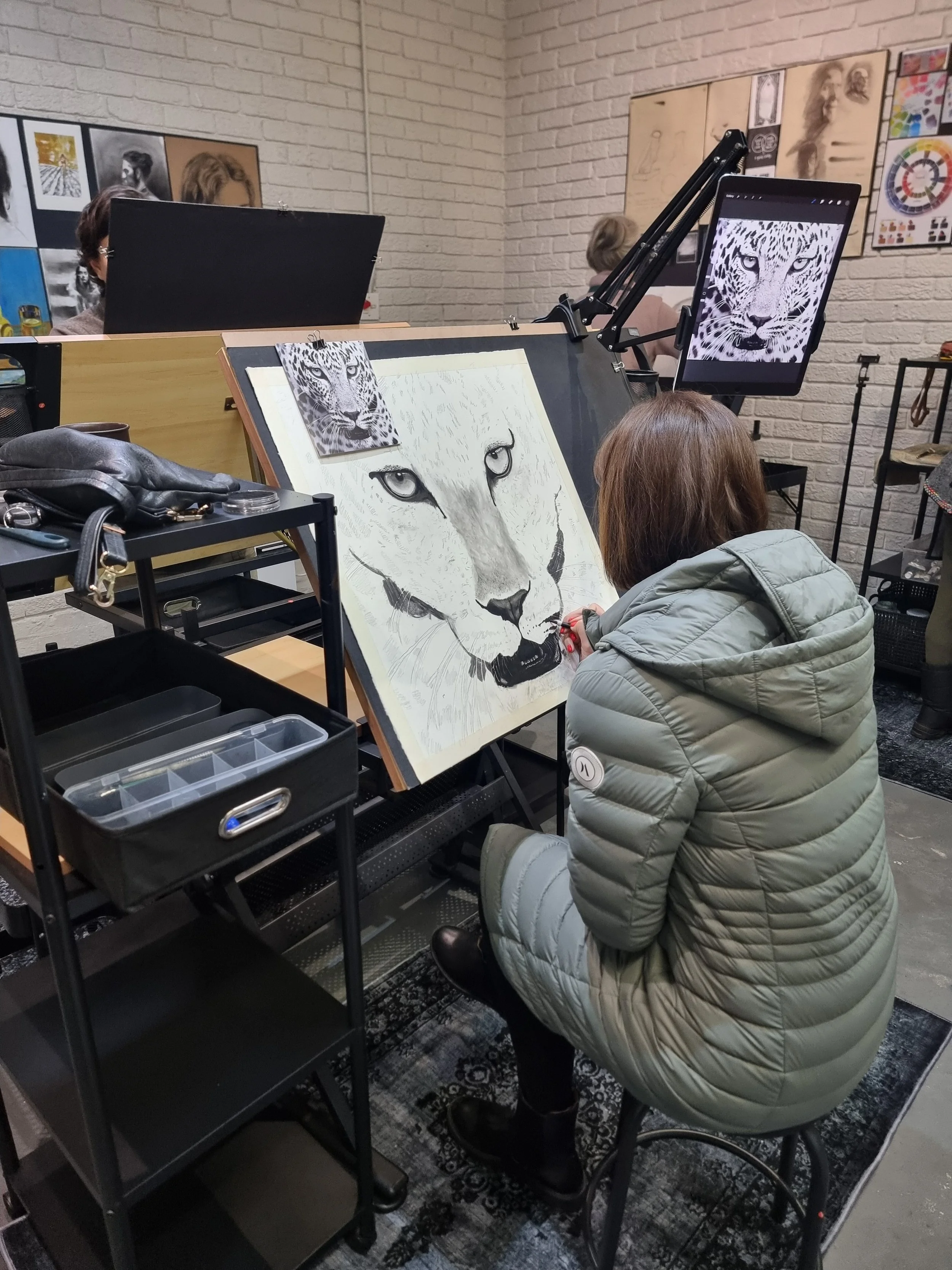 A woman in a gray jacket sketches a large realistic pencil drawing of a white tiger's face on an easel, with sample images of the tiger's face attached to the workspace, in an art studio with other artwork .Art lessons for adults Durbanville
