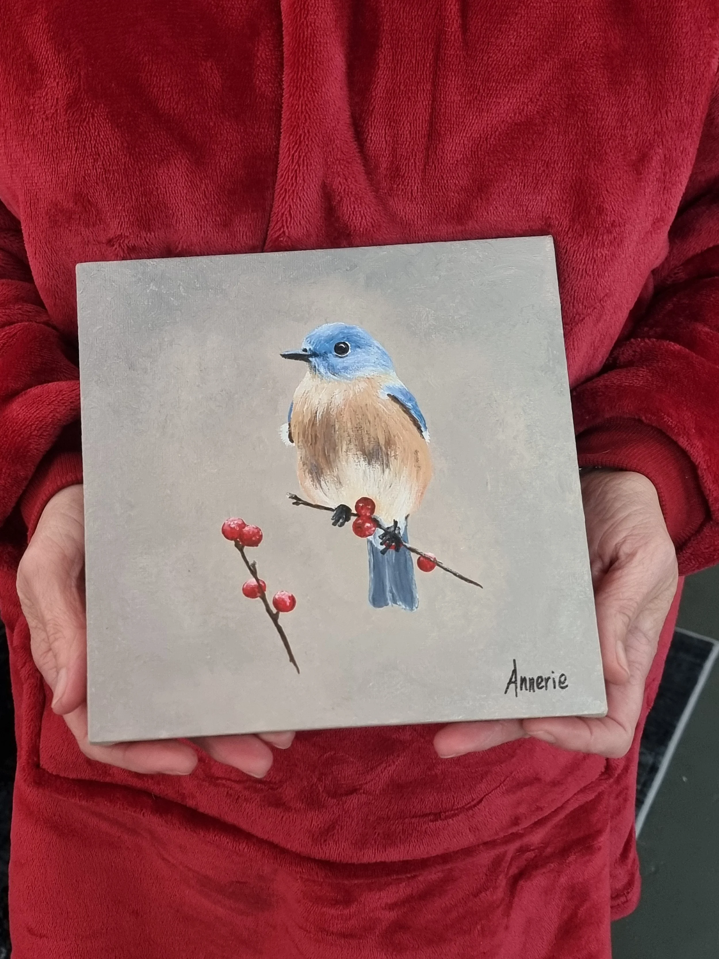 Person wearing a red jacket holding a canvas painting of a bird with blue head and brown body perched on a branch with red berries. this is Beginner art classes Durbanville