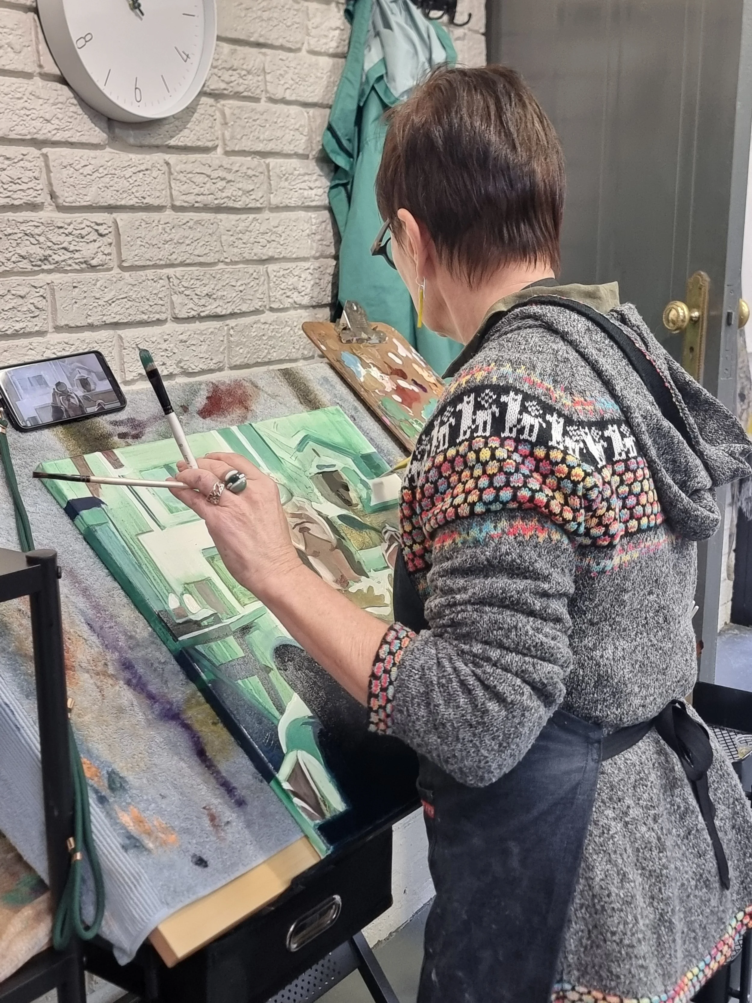 A woman painting a landscape on a canvas, with a smartphone propped up beside her showing her live video of herself painting, in an art studio with a brick wall and clock in the background.