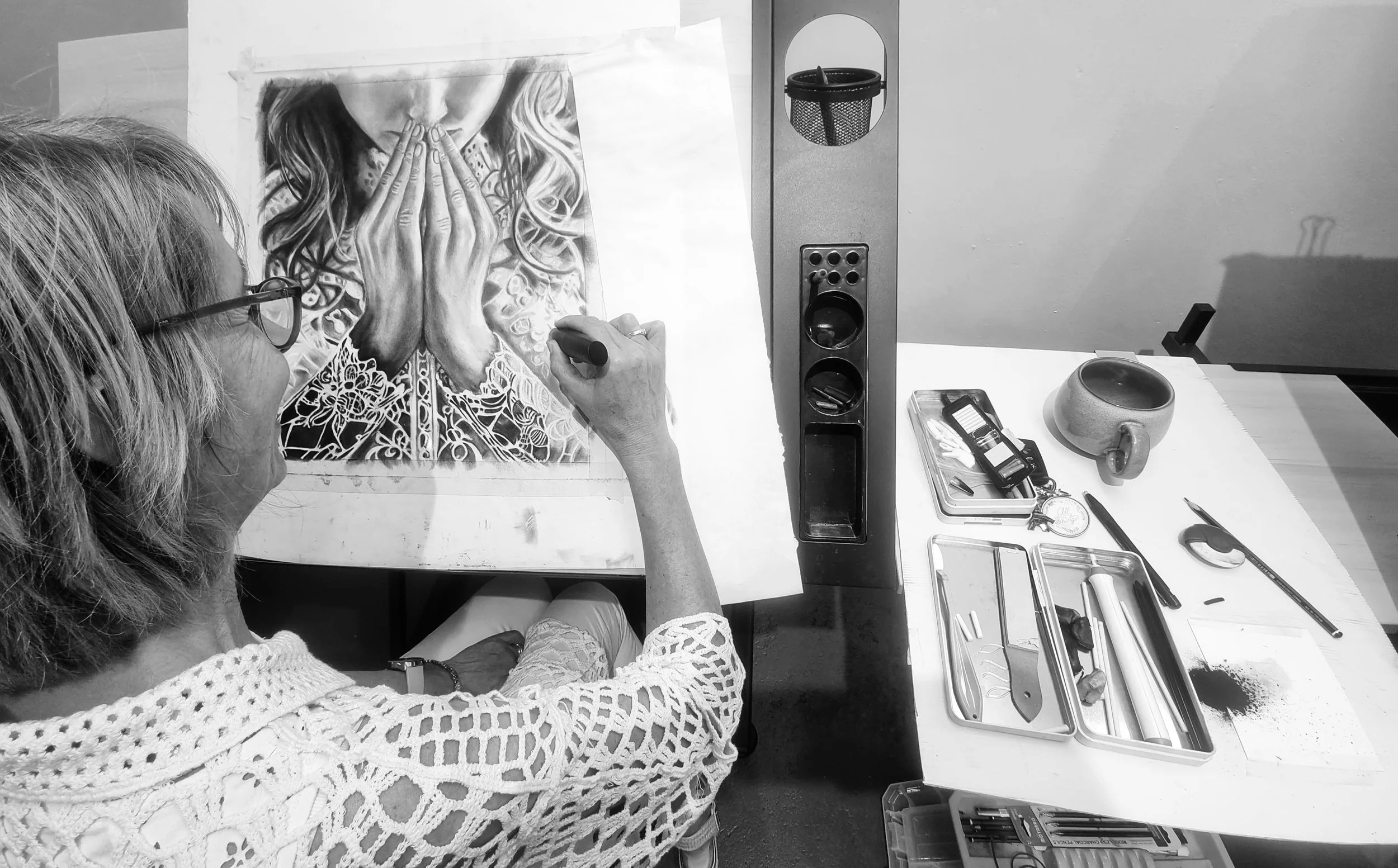 An artist working on a detailed black and white portrait of a woman with her hands covering her mouth, at an art station with various drawing tools and supplies. Adult beginner art classes near me